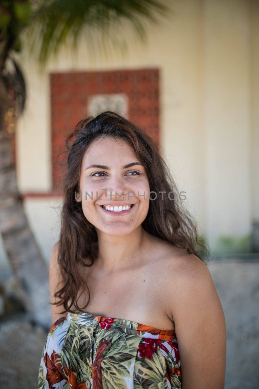 Beautiful woman wearing flowered dress standing in a tropical street with palm trees as background. Portrait of smiling brunette woman on sunny day. Tropical summer vacations