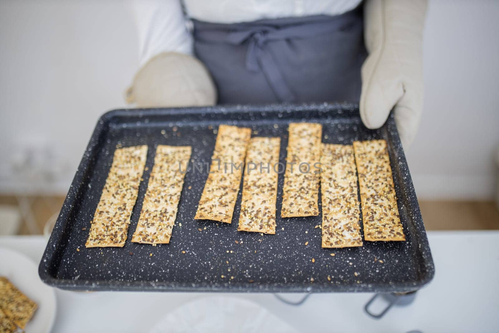 Gloved hands holding black tray of Swedish multi-seed crispbread. Freshly baked crispy bread strips with linseed, amaranth, and rye. Healthy baked snacks