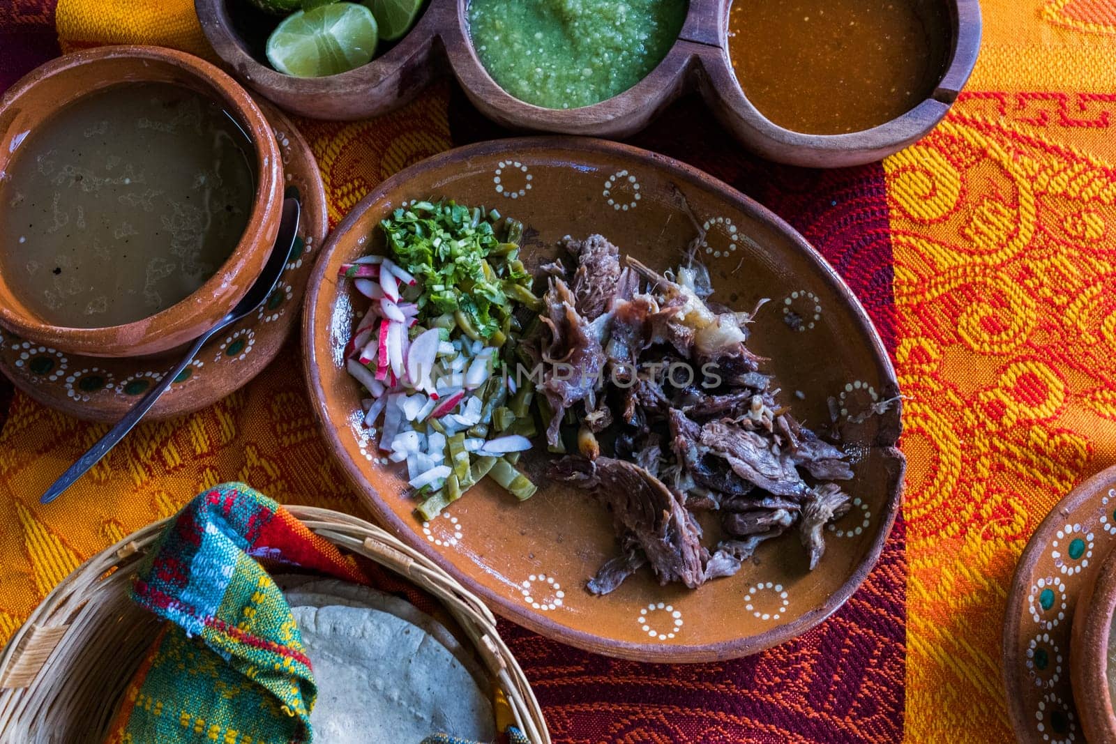 Top view of chopped lamb meat, hot sauces, lime slices, broth, and tortillas on colorful tablecloth. Tasty meal and condiments in clay plates and baskets above red and orange table. Mexican cuisine
