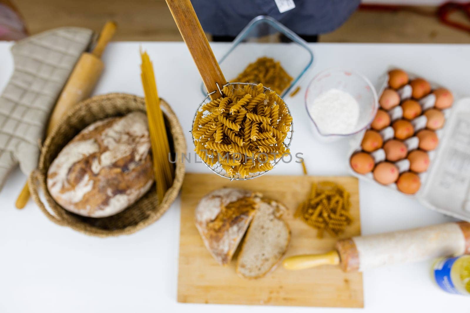 Uncooked fusilli on wooden pasta strainer above ingredients on white table. Eggs, spaghetti, flour, and bread on table from above. Pasta preparation