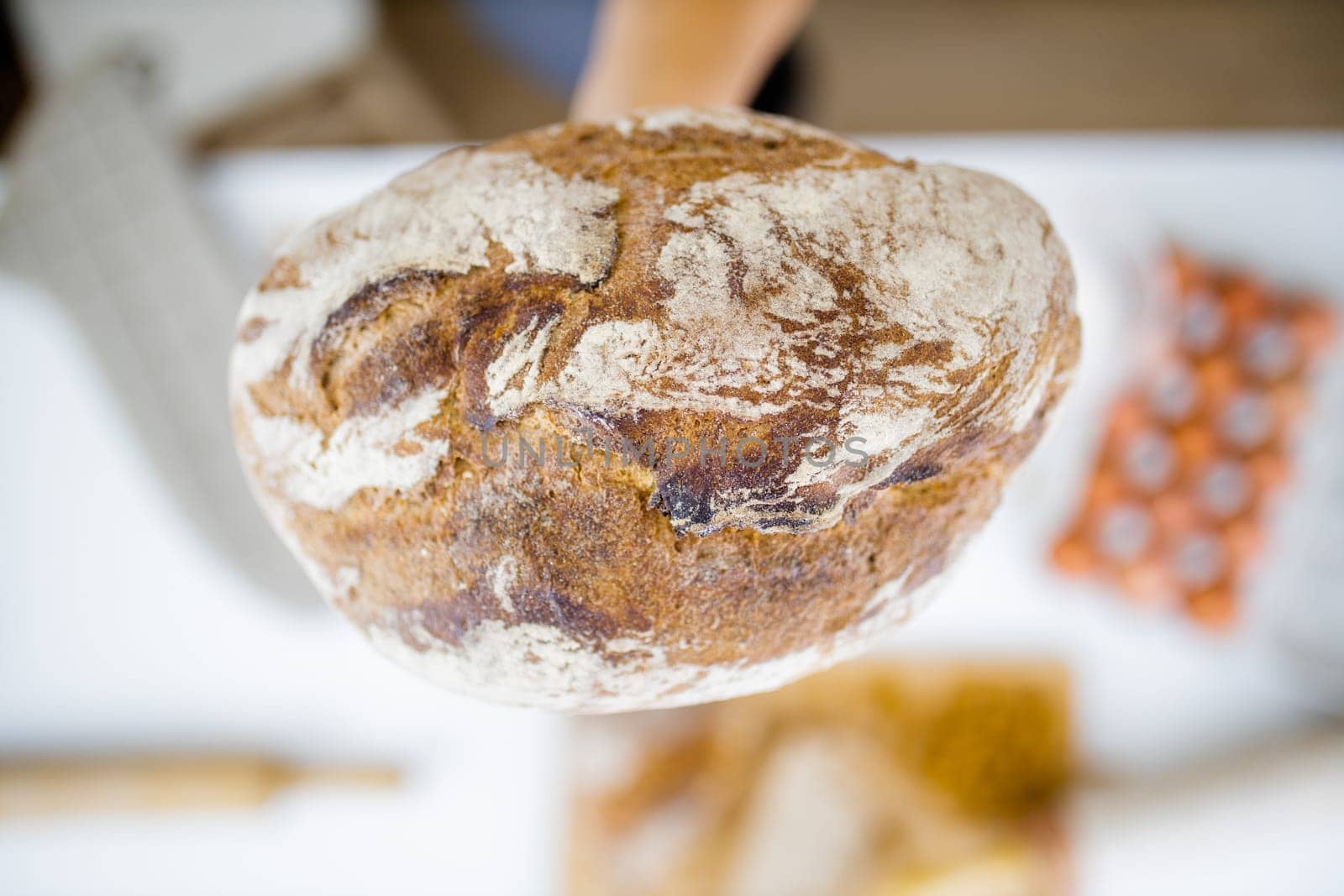 Female hand holding loaf of bread above blurry ingredients on table. Hand holding sourdough bread from above. Healthy meal preparation