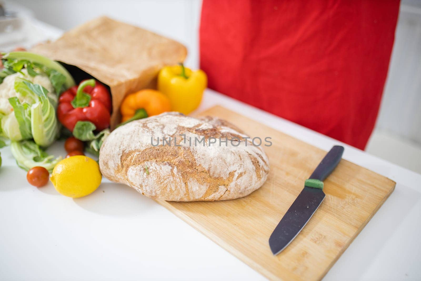 Bread on cutting board above white table and next to paper bag of vegetables. Sourdough bread alongside bell peppers, cauliflower, and lemons. Healthy meal preparation
