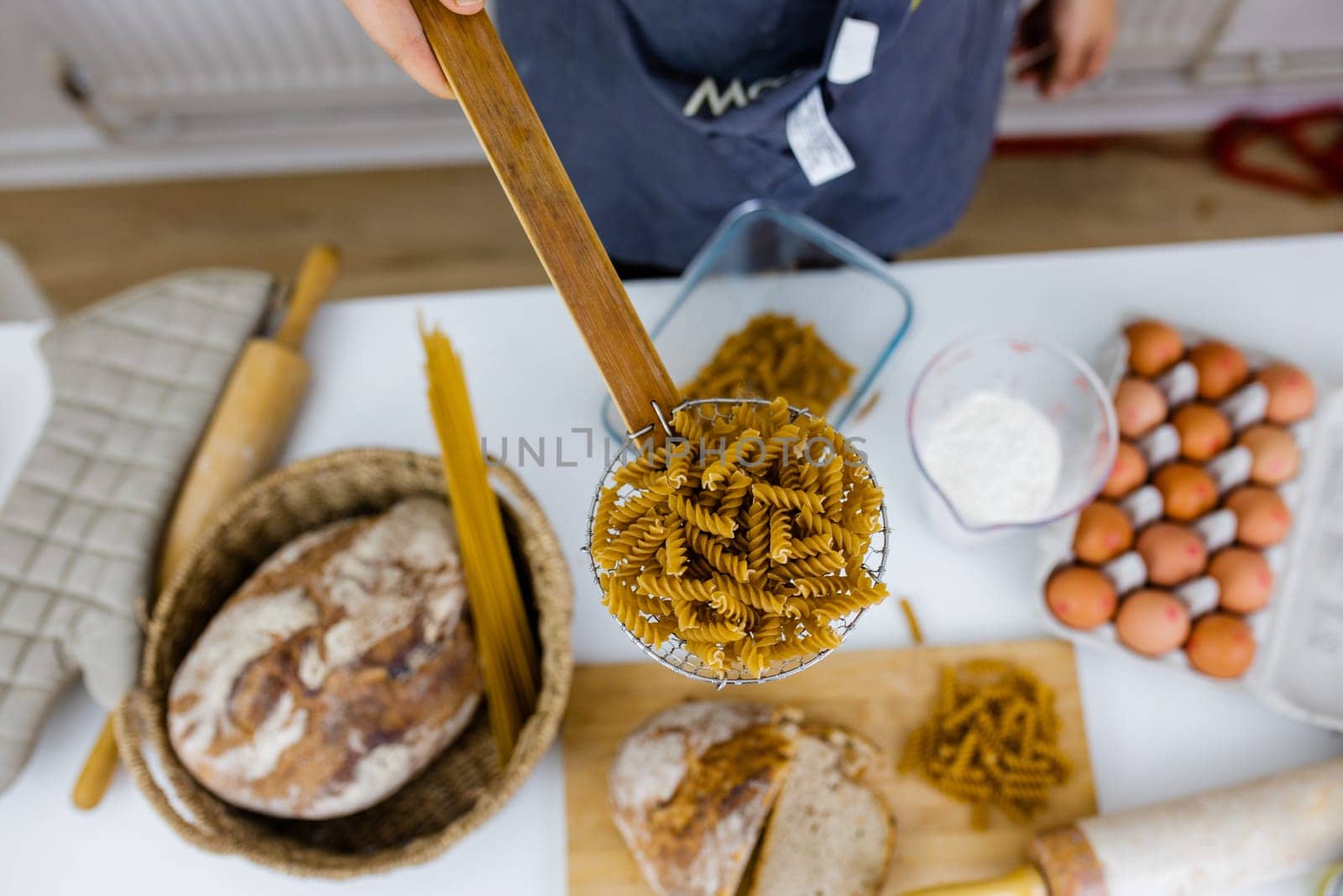 Uncooked fusilli on wooden pasta strainer above ingredients on white table. Eggs, spaghetti, flour, and bread on table from above. Pasta preparation