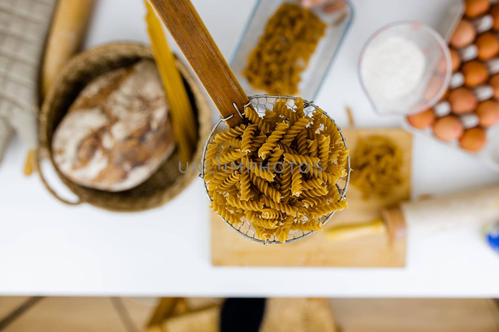 Uncooked fusilli on wooden pasta strainer above blurry ingredients on white table. Blurry eggs, spaghetti, flour, and bread on table from above. Pasta preparation