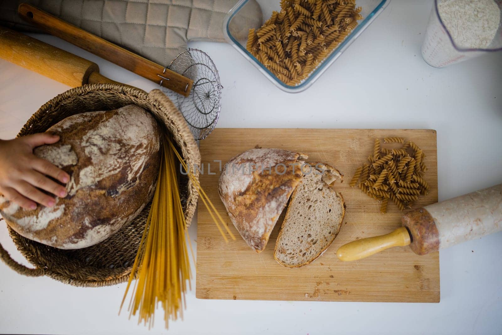 Bread, pasta, flour, and wooden cookware on white table from above. Ingredients and carbohydrates on cutting board and a basket. Healthy meal preparation