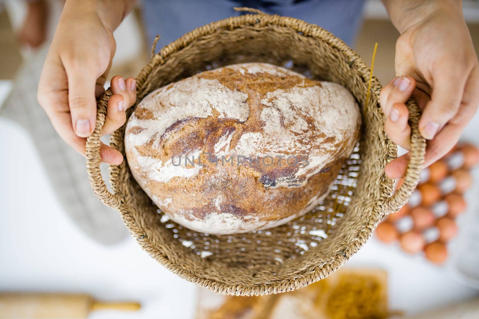 Female hands holding basket of sourdough bread above blurry ingredients. Blurry eggs, pasta, flour, and bread on table from above. Healthy meal preparation