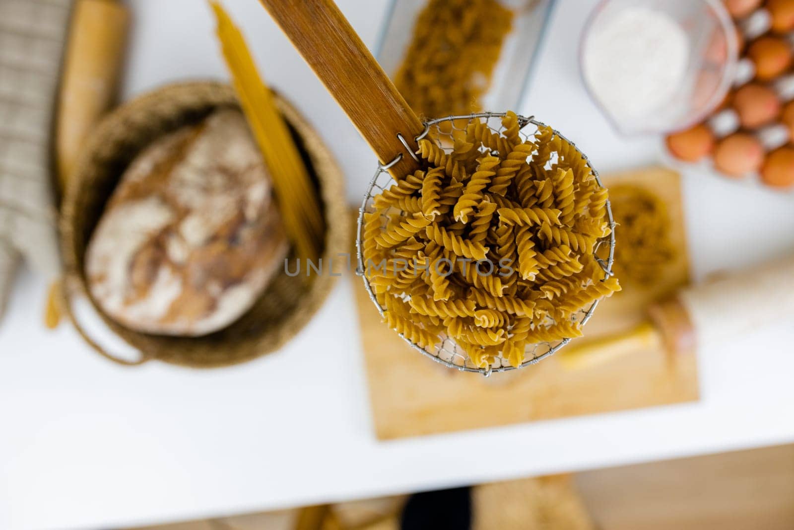 Uncooked fusilli on wooden pasta strainer above blurry ingredients on white table. Blurry eggs, spaghetti, flour, and bread on table from above. Pasta preparation