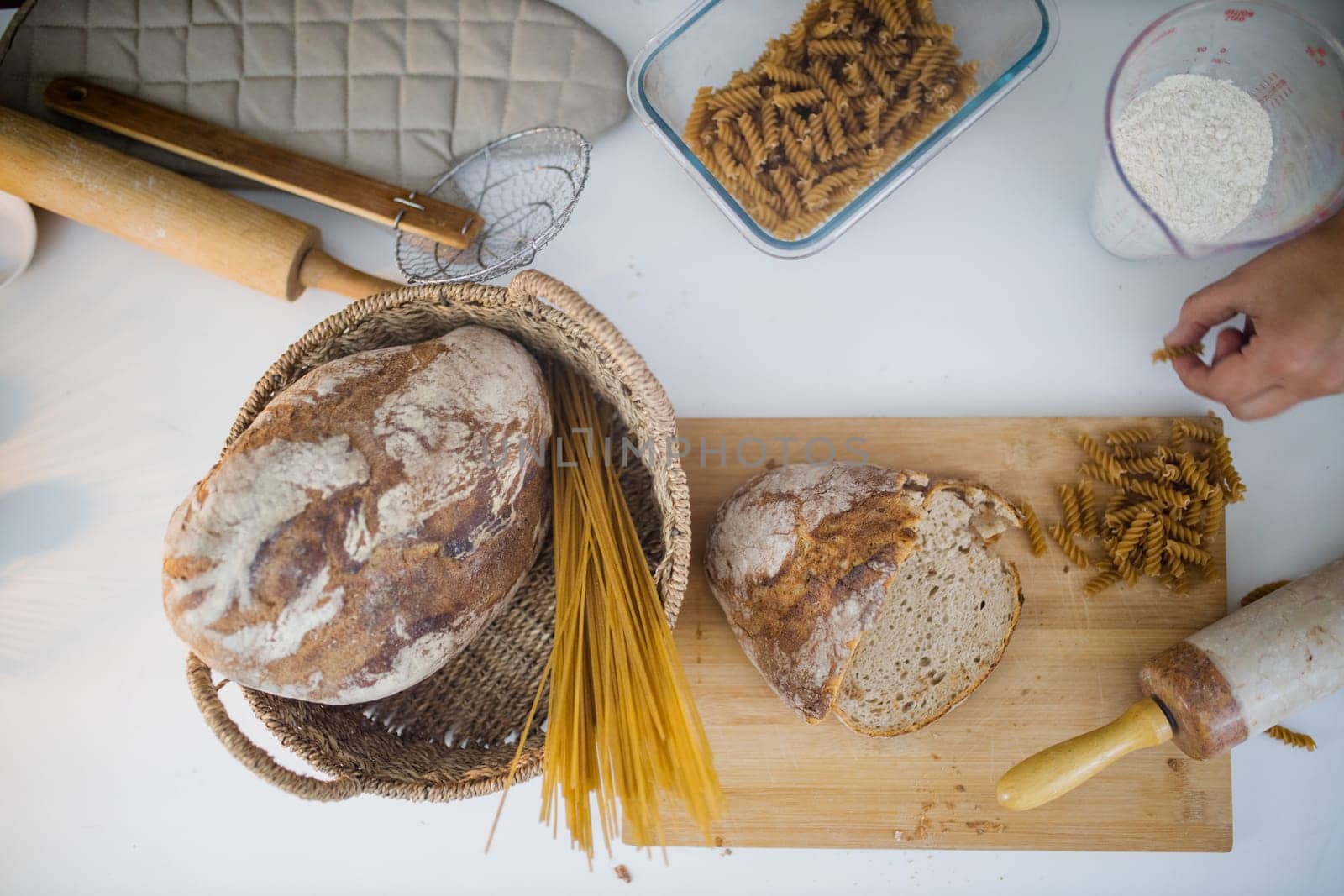 Bread, pasta, flour, and wooden cookware on white table from above. Ingredients and carbohydrates on cutting board and a basket. Healthy meal preparation