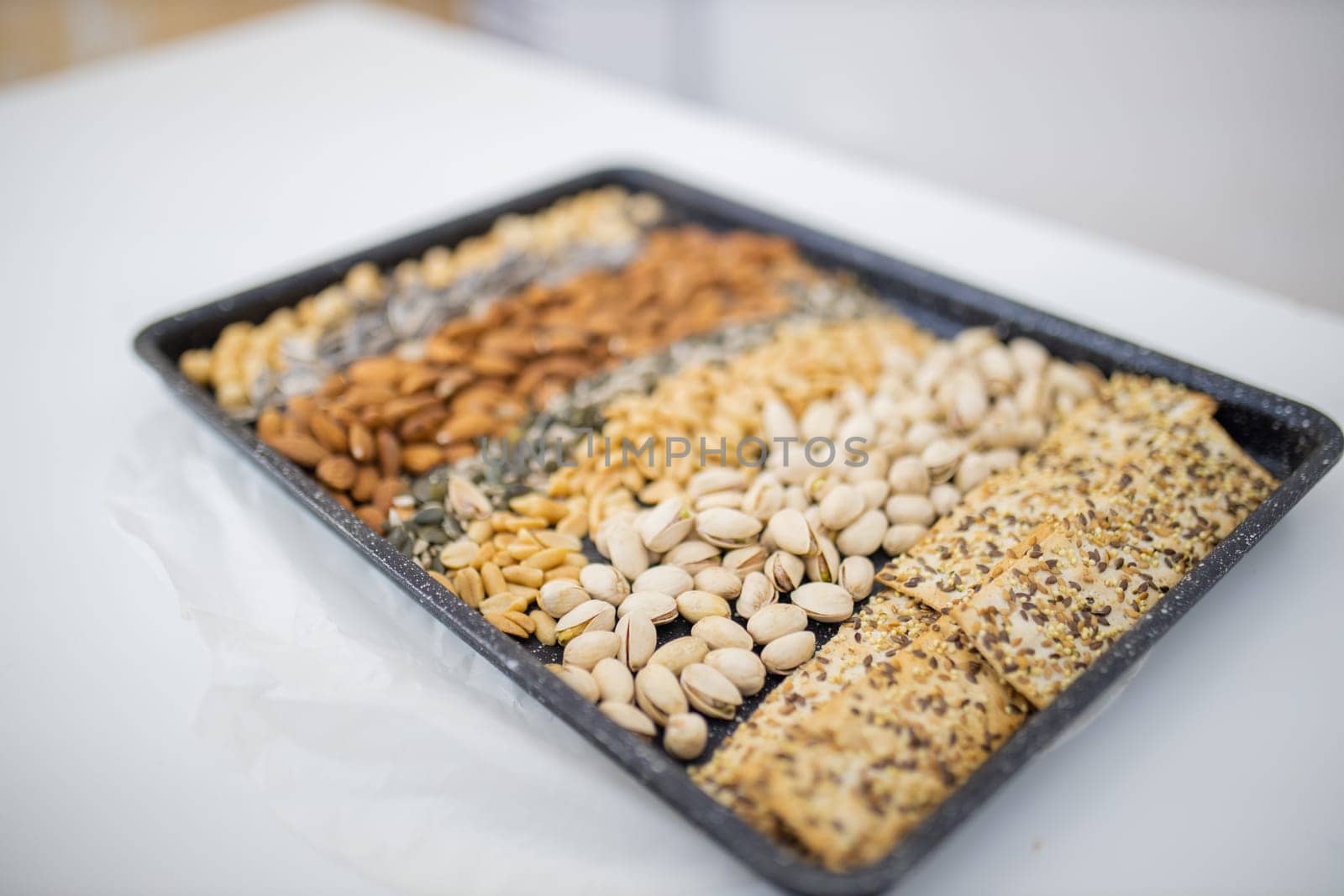Black tray of chickpeas, almonds, peanuts, pistachios, and multi-seed crispbread. Fresh crispy bread, seeds, and nuts above white table. Healthy snacks