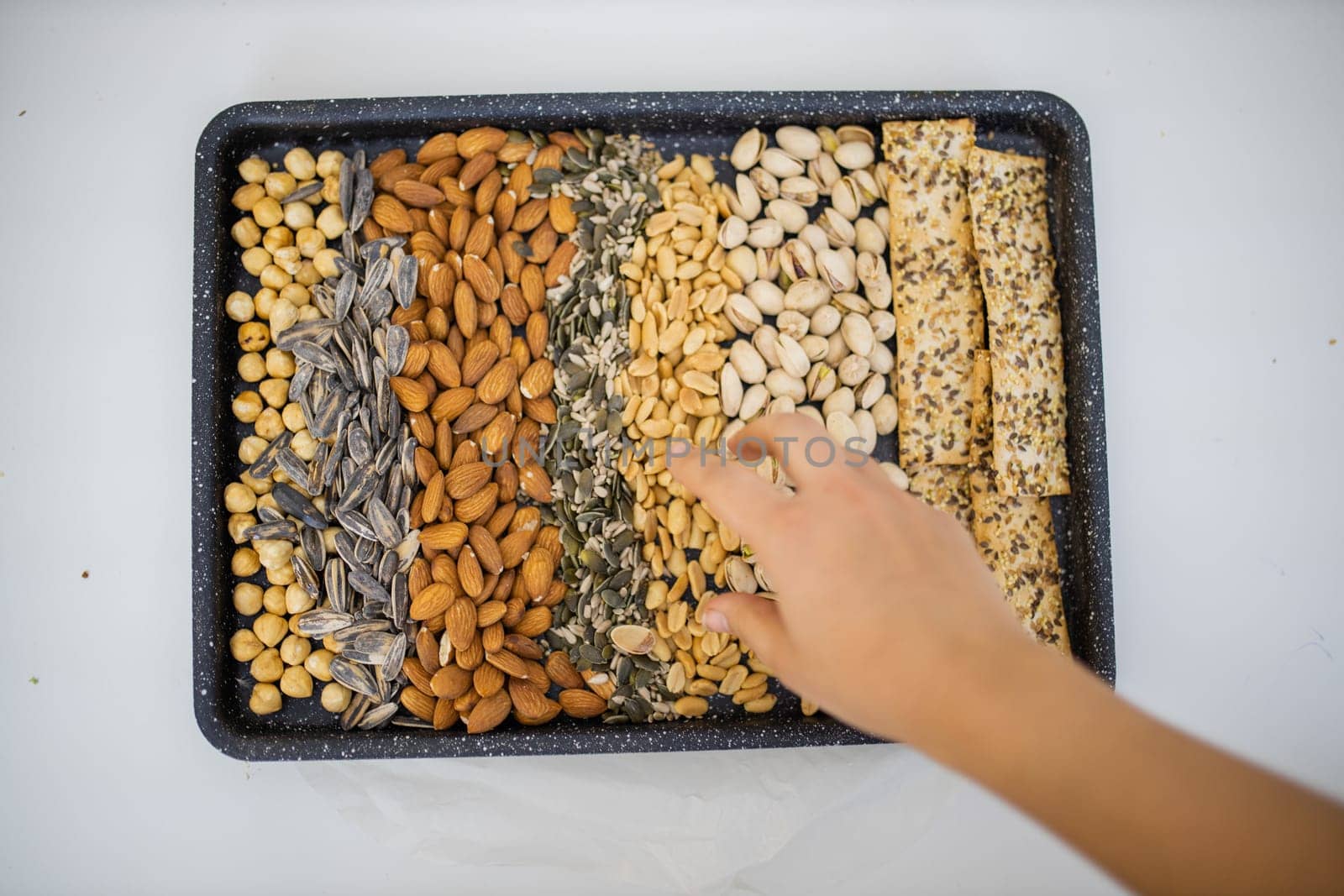 Female hand picking peanuts from black tray of chickpeas, almonds, pistachios, and multi-seed crispbread. Fresh crispy bread, seeds, and nuts from above. Healthy snacks