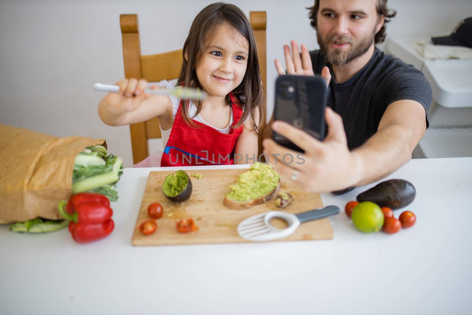 Happy father and daughter taking selfie while spreading avocado on toast. Cute young child cutting vegetables on cutting board. Father-daughter cooking together