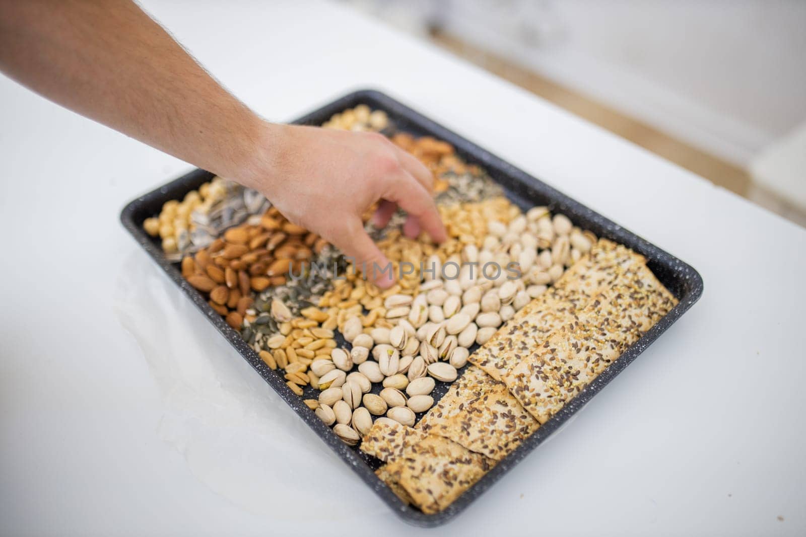 Male hand picking peanuts from black tray of chickpeas, almonds, pistachios, and multi-seed crispbread. Fresh crispy bread, seeds, and nuts above white table. Healthy snacks