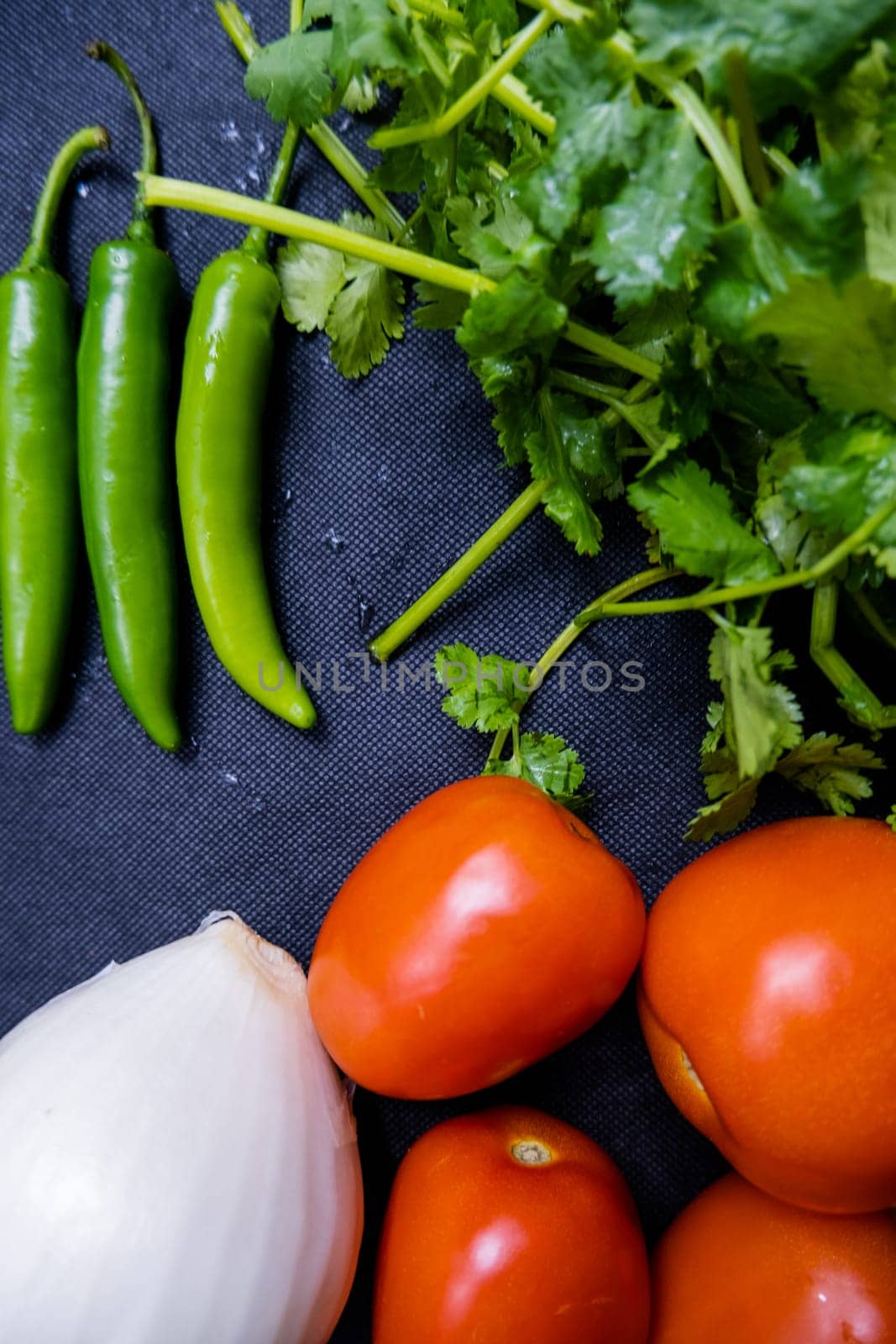 Top view of three green chili peppers, onion, four tomatoes, and coriander on table. Fresh and aromatic vegetables and herbs on dark blue surface. Healthy meal preparation