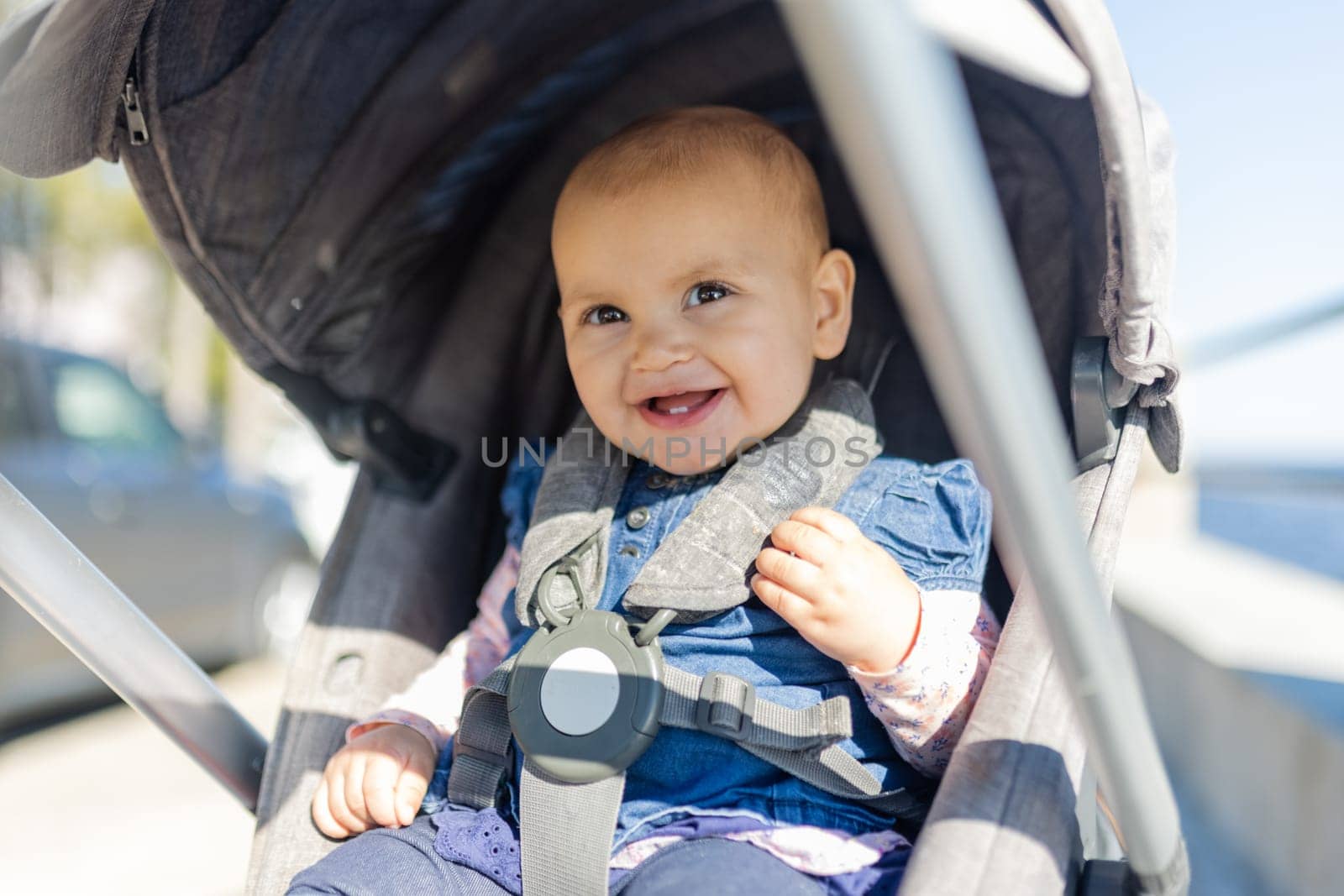 Adorable view of bright sunlight on happy baby sitting in gray stroller with blurry background. Portrait of cute smiling infant in blue dress during sunny day. Peaceful toddlers outdoors