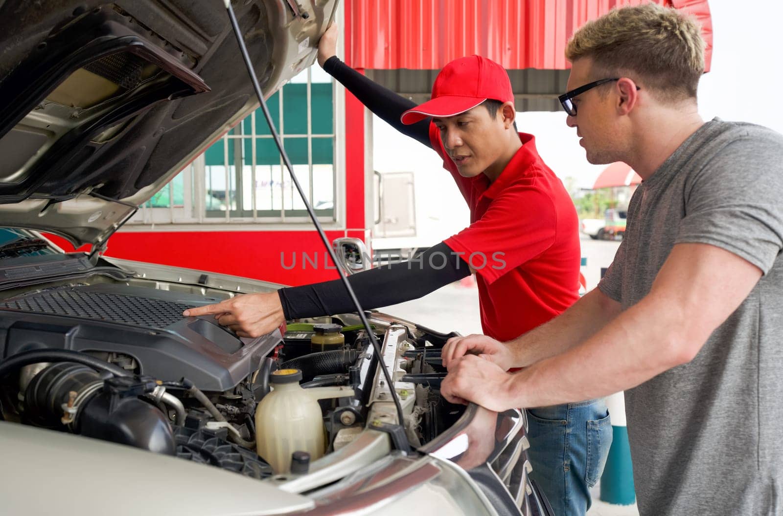 An Asian gas station worker raises the car bonnet to check for the cause of an engine malfunction. A caucasian driver with sunglass stand listening attentively in front of broken pickup truck.