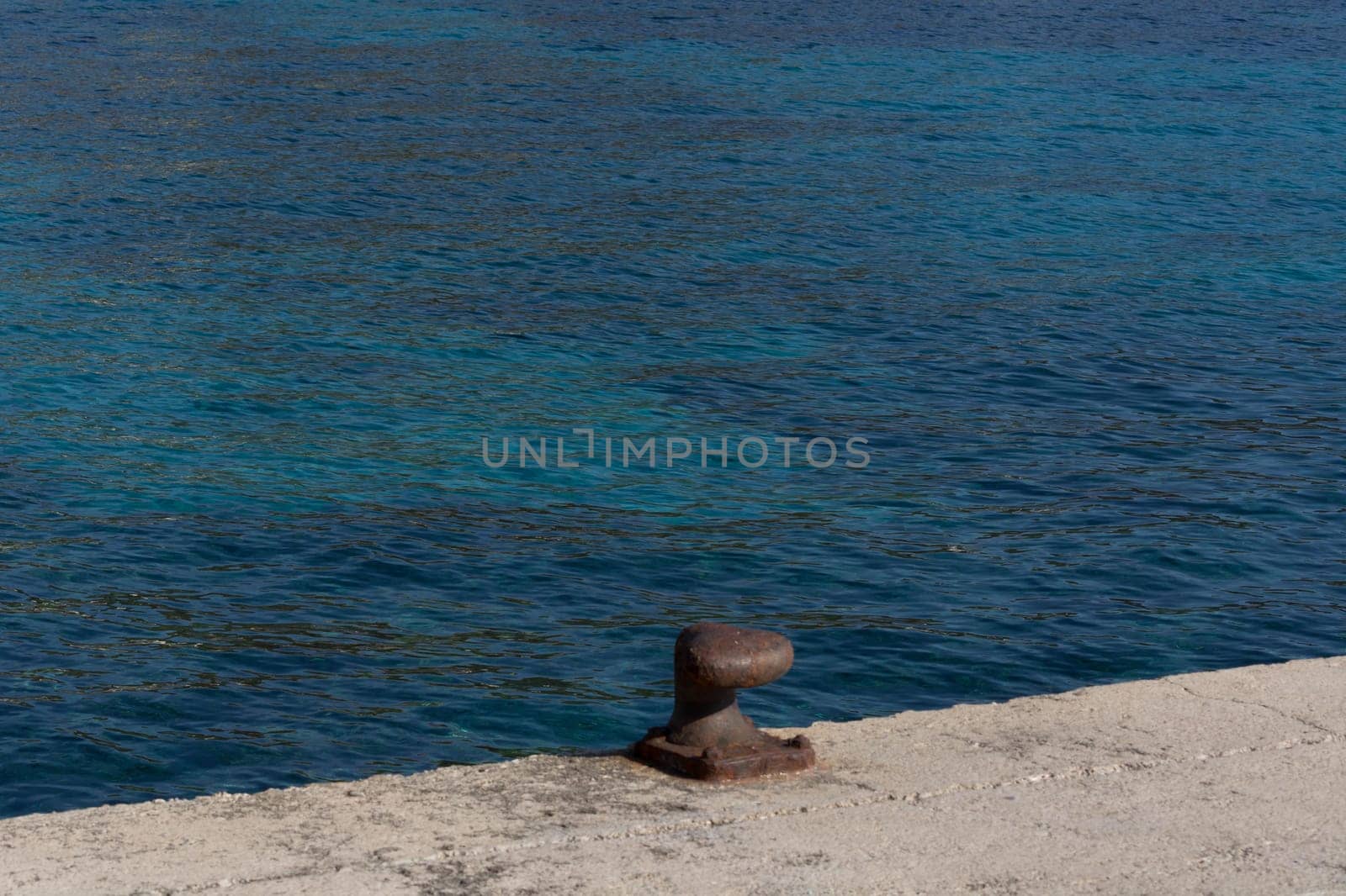 A rusty bollard on a quay wall with a rusty chain and in the background the water in the harbor on the Mediterranean Sea.