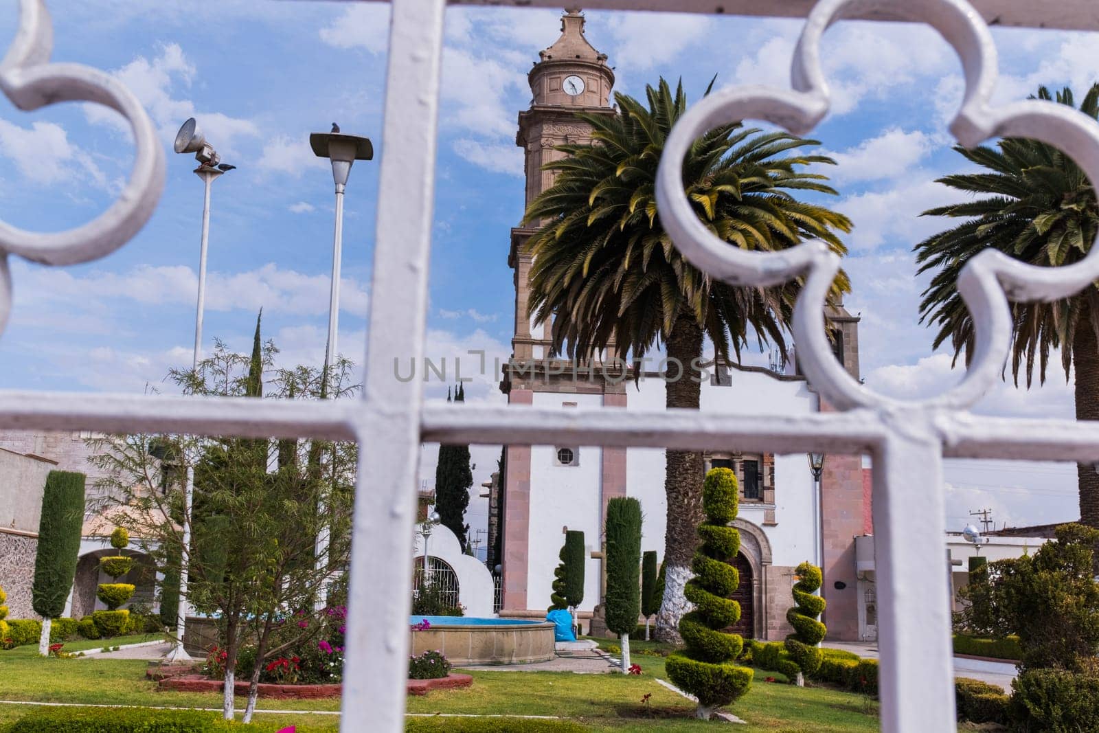 Beautiful view from behind white metal fence of old Hispanic church surrounded by bushes and palm trees. Ancient bell tower with slightly cloudy sky as background. Classic Mexican architecture