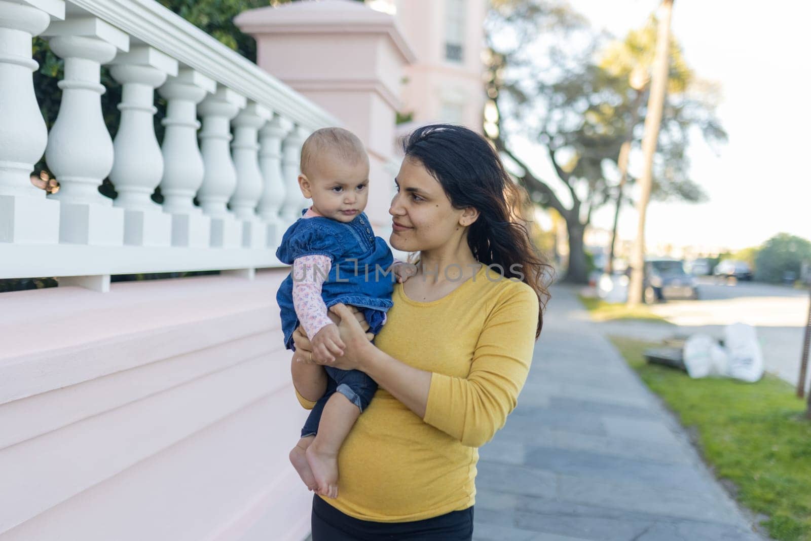 Portrait of happy mother holding her cute baby outside white and pastel-pink building. Beautiful brunette woman and her adorable young daughter in peaceful neighborhood. Lovely family outdoors