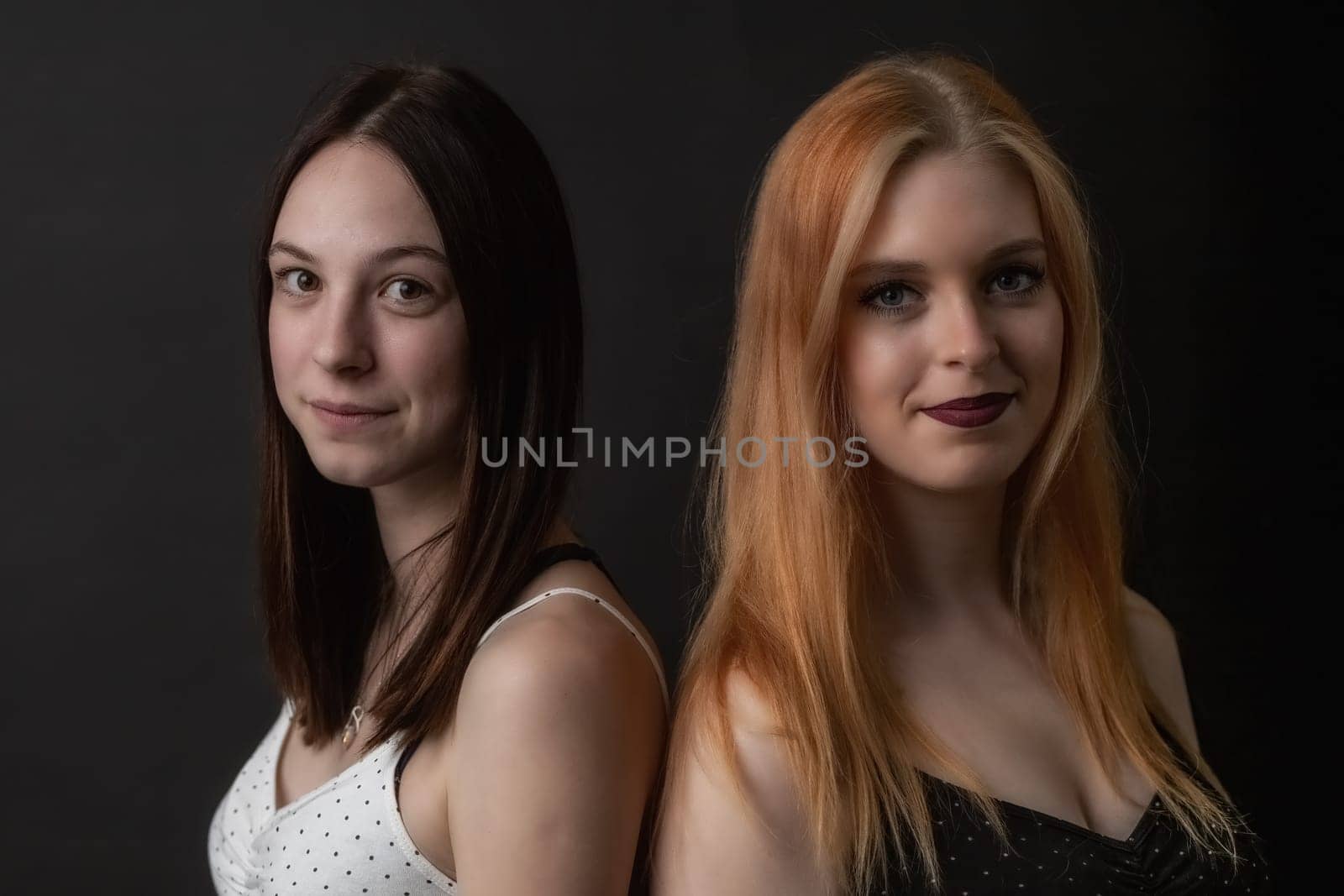 Pair of smiling young women is posing on the black background. Horizontally.