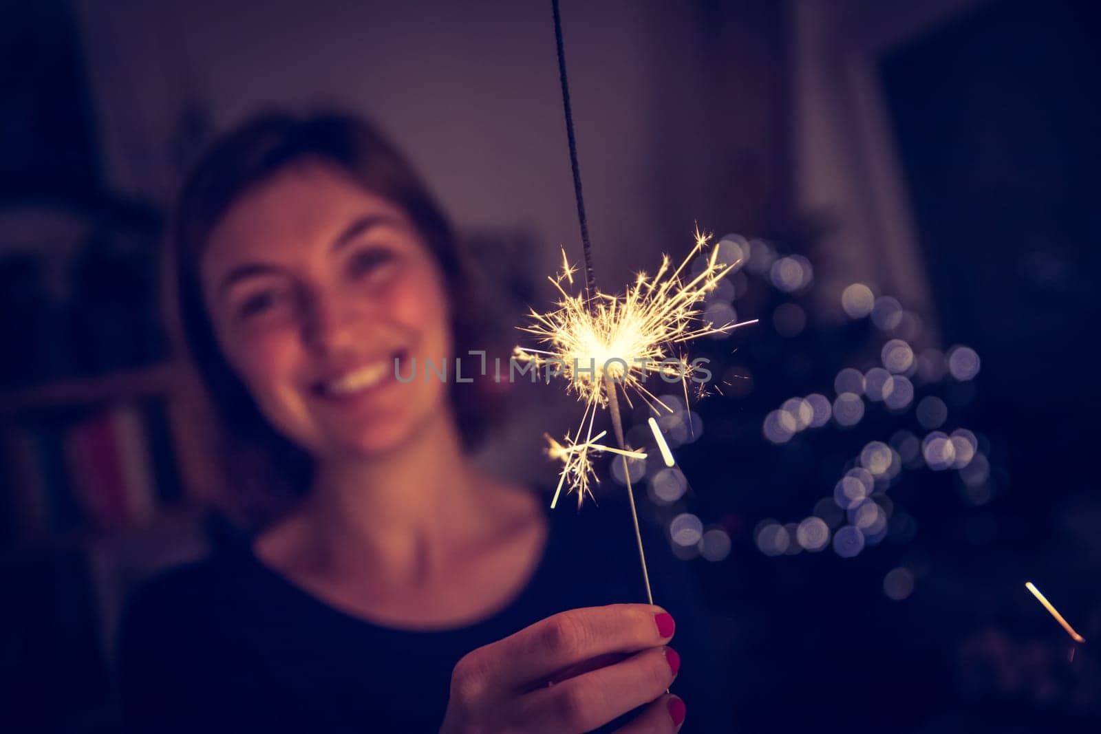 Smiling girl is holding a sparkler in her hand, indoor, lights in background