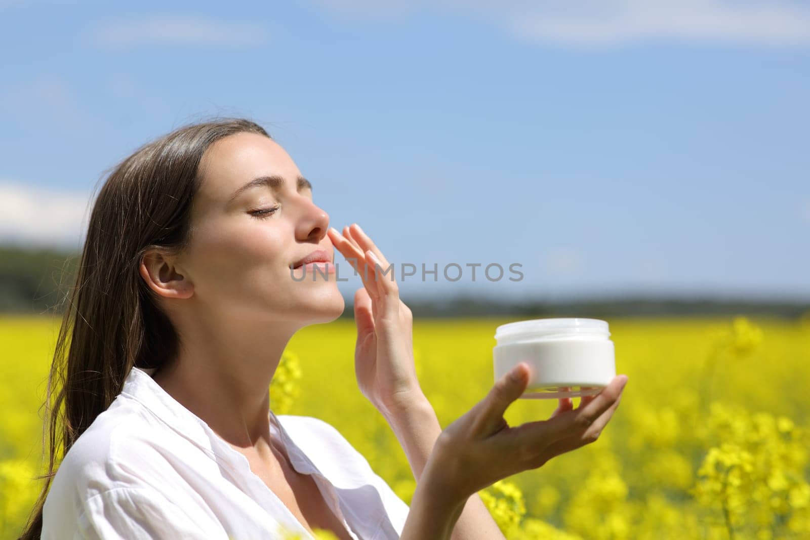 Beauty woman holding jar applying moisturizer cream on face in a flowered field