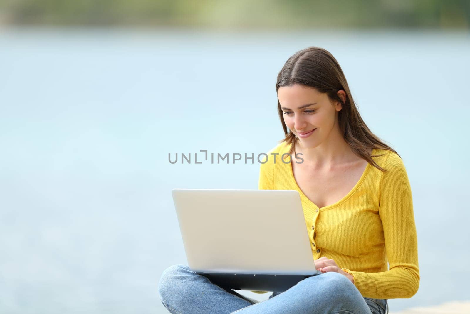 Happy woman using a laptop sitting beside a lake