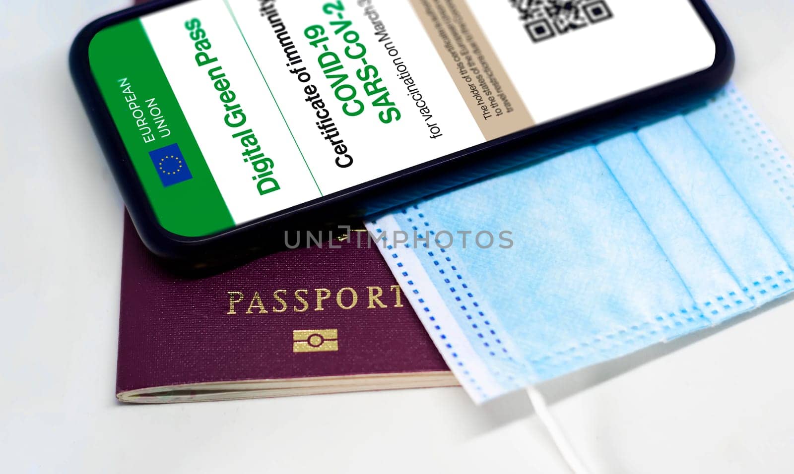 The digital green pass of the european union with the QR code on the screen of a mobile phone over a surgical mask and a passport. Immunity from Covid-19. Travel without restrictions.