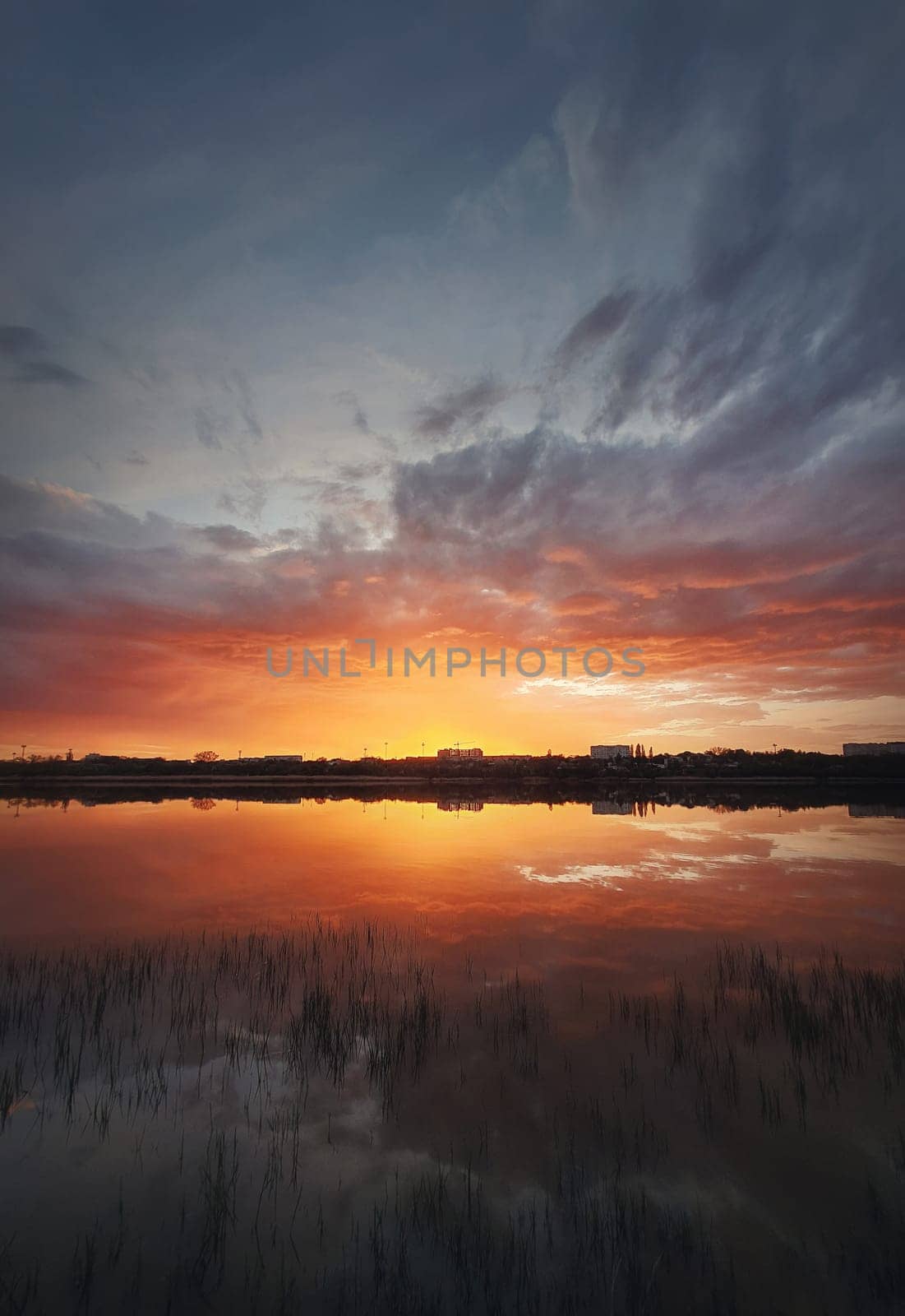 Fascinating sunset reflecting on the lake surface. Idyllic landscape, vertical background. Silent and tranquil evening scene with colorful sky, reed vegetation in the pond and a city at the horizon