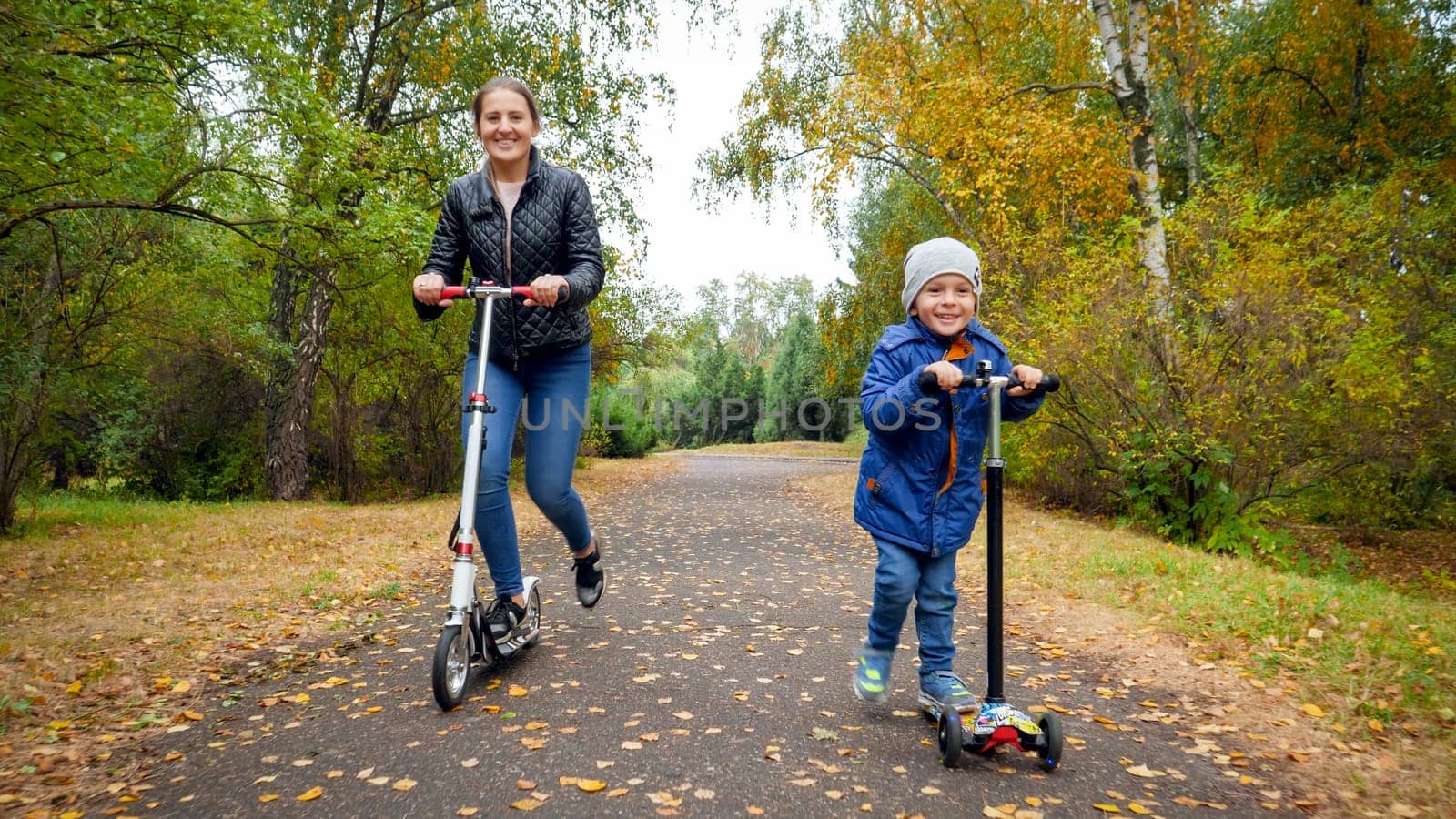 Happy smiling boy with mother racing on kick scooters at autumn park.