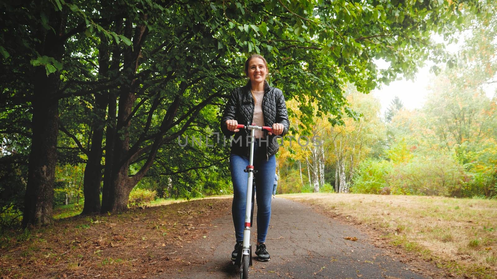 Beautiful smiling young woman riding on kick scooter at autumn park.
