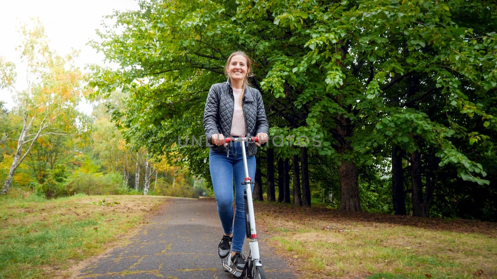 Happy young woman riding on scooter under big trees at autumn park.