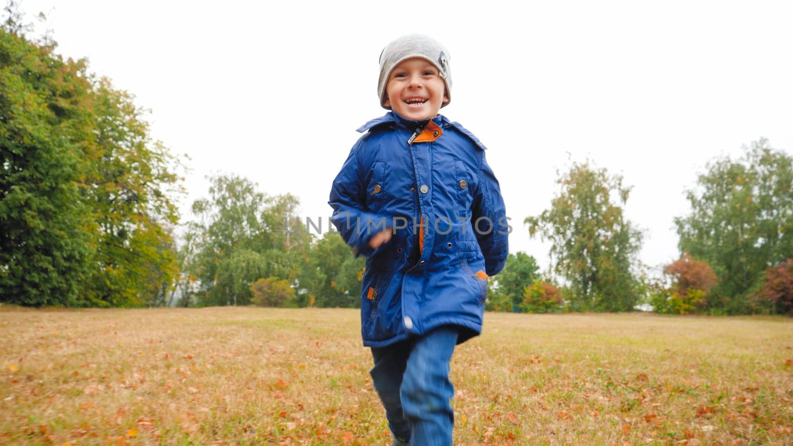 Portrait of laughing little boy running on grass at field on cloudy autumn day.