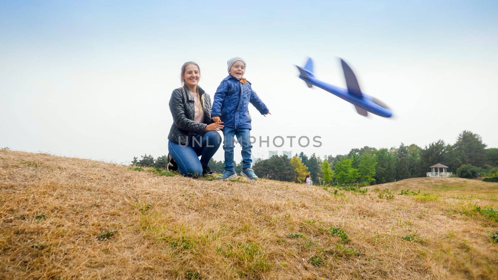 Happy excited little boy with mother throwing toy airplane from the hill at park.