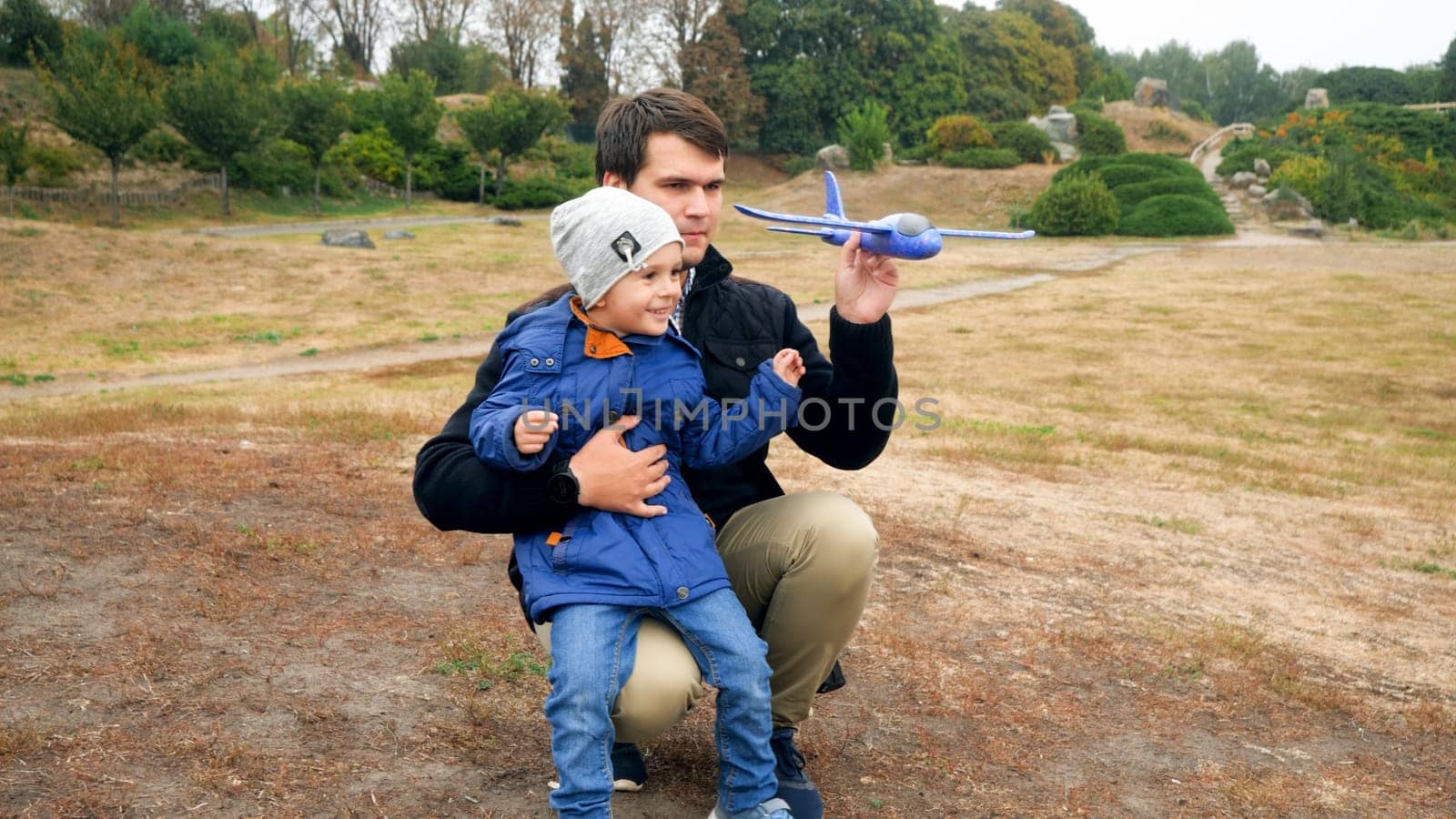 Little smiling boy with young father playing with toy airplane in the field.