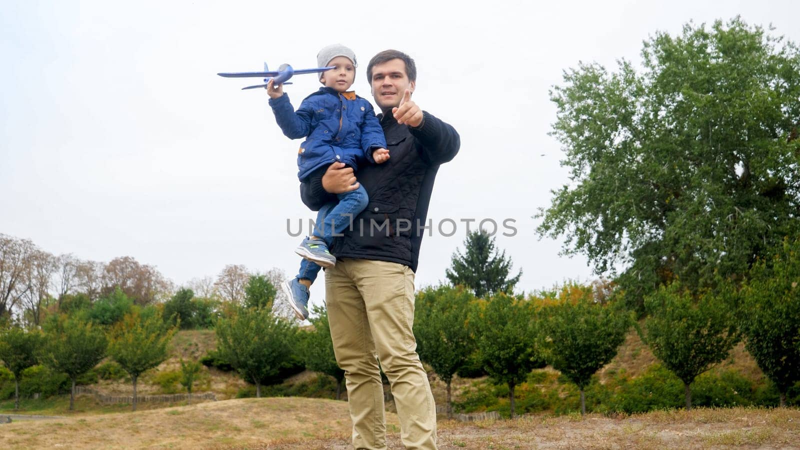 Portrait of young father holding his little son and teaching throwing toy airplane at park.