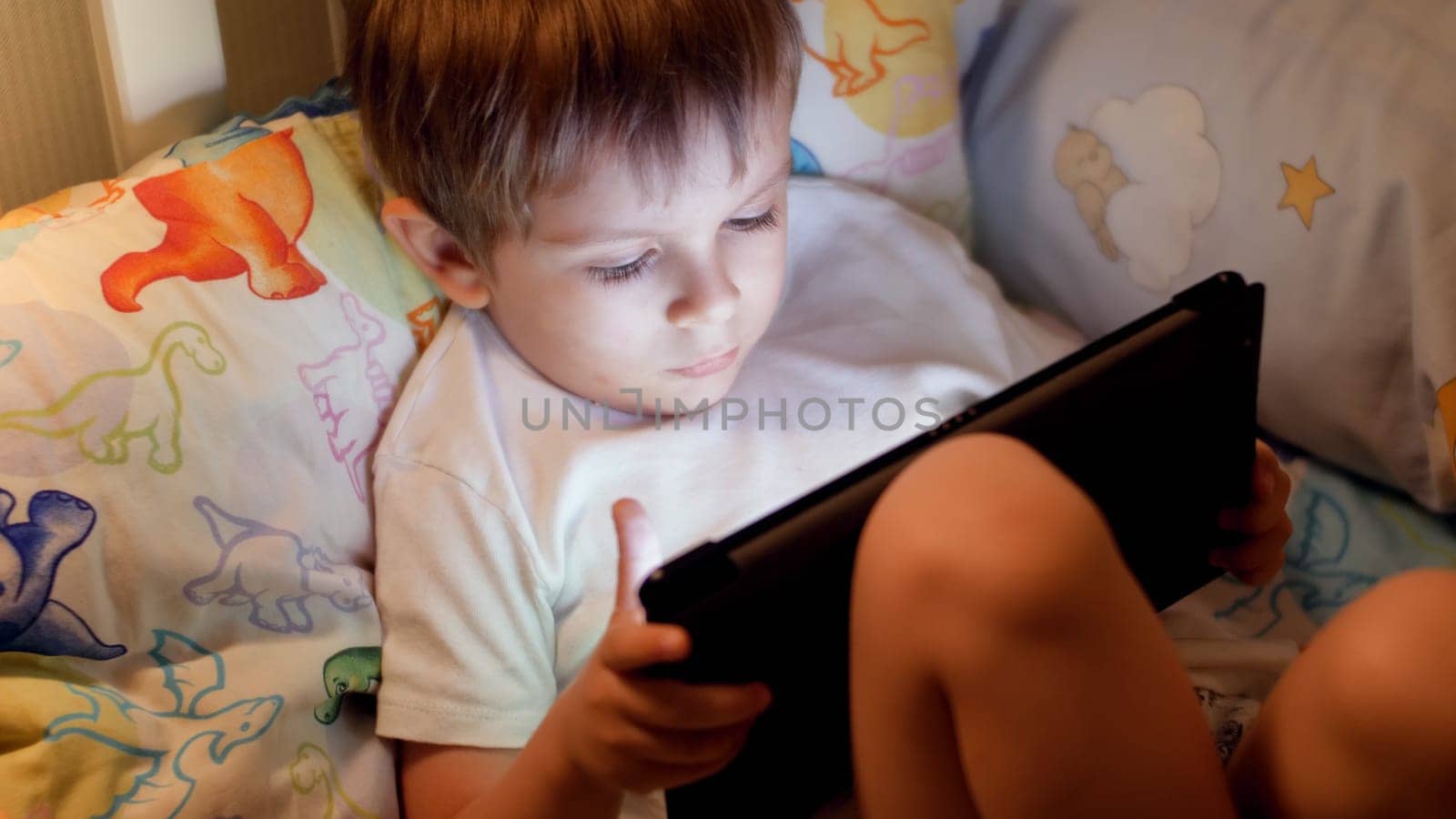 Closeup photo of little boy going to sleep and browsing internet on digital tablet computer.