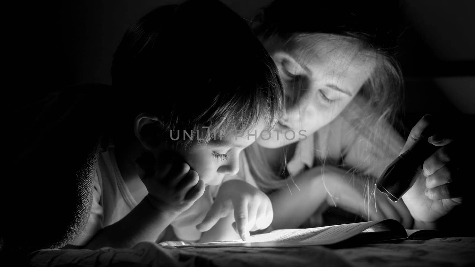 Black and white portrait of little toddler boy reading book with mother lying under blanket at night.