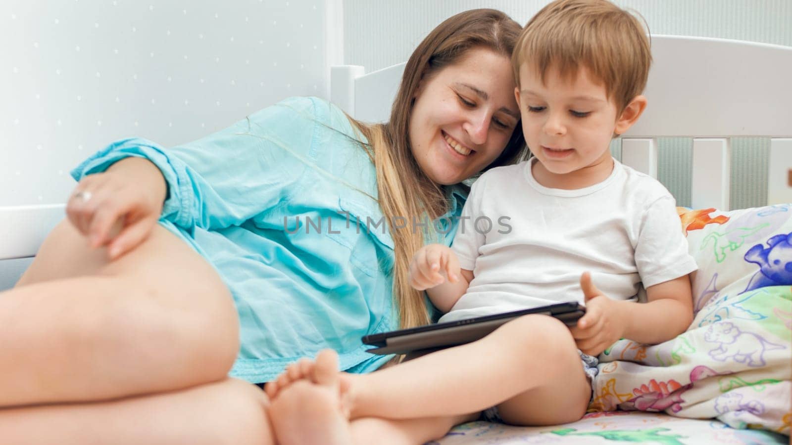 Portrait of smiling young mother with little son lying in bed and using digital tablet computer.