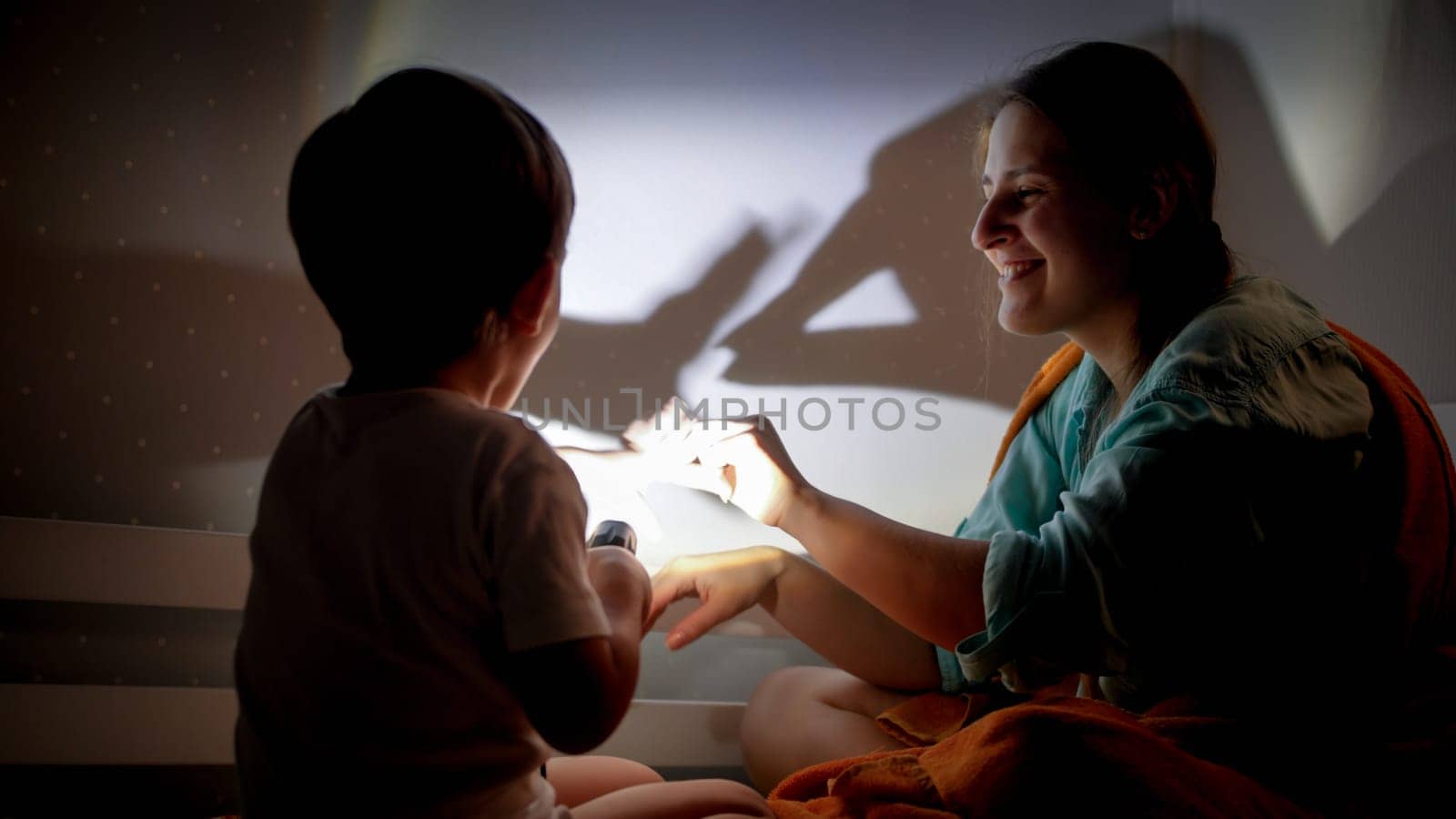 Portrait of smiling young mother and little boy playing with flashlight and shadows on wall at night.