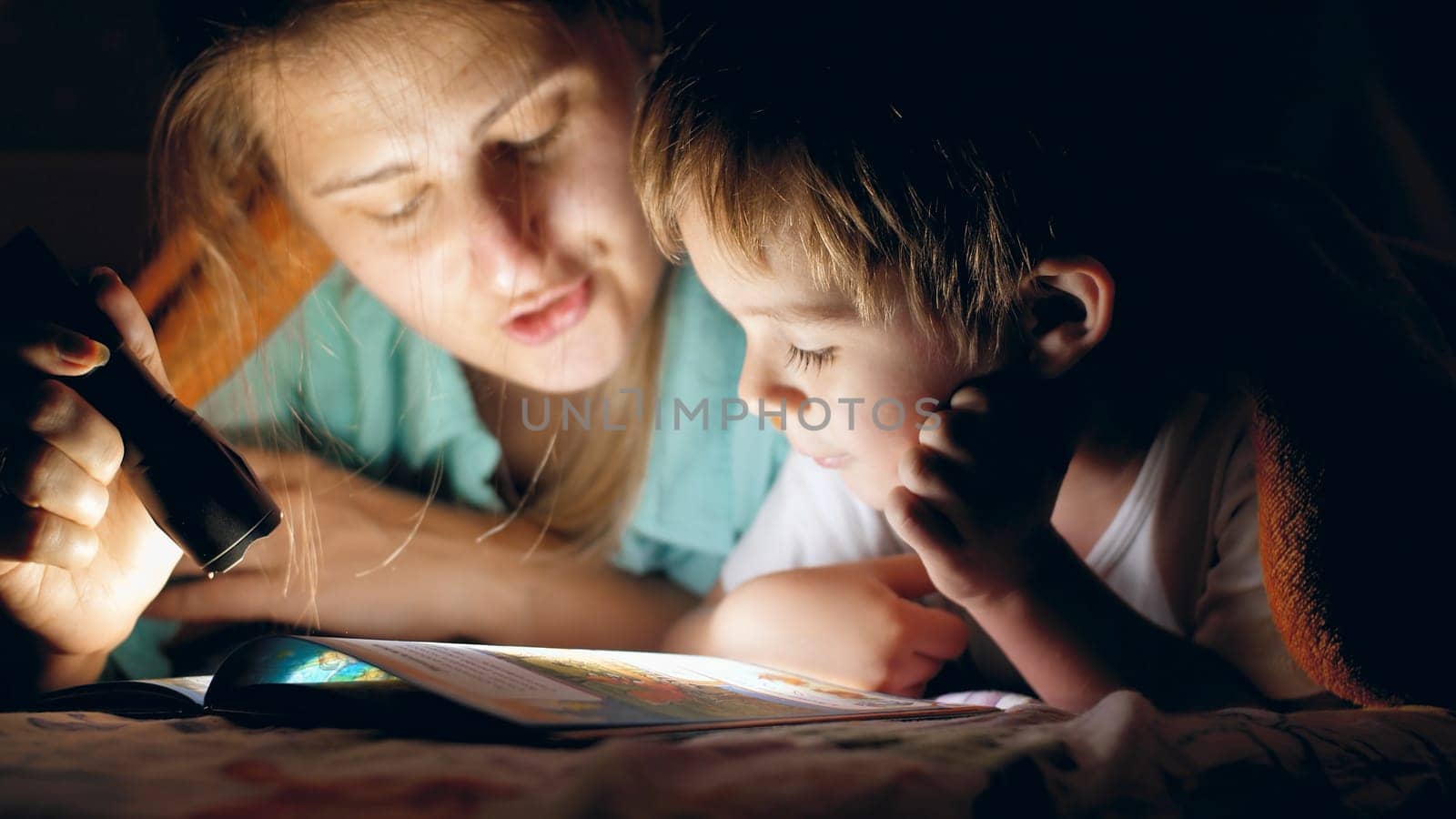 Toned portrait of young mother holding flashlight and reading bedtime story book to her little son lying under blanket at night.