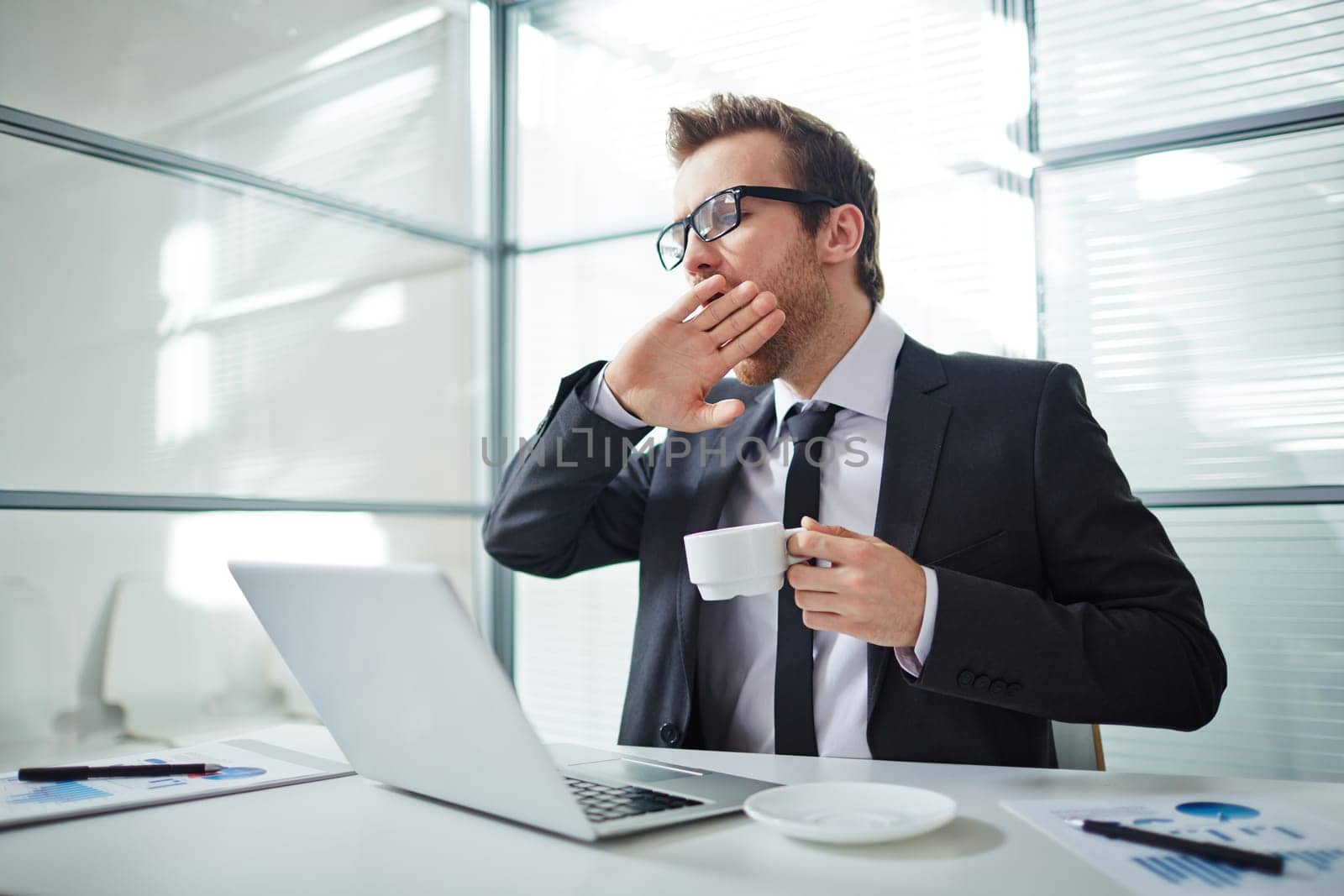 Tired man with cup of coffee yawning at workplace in office