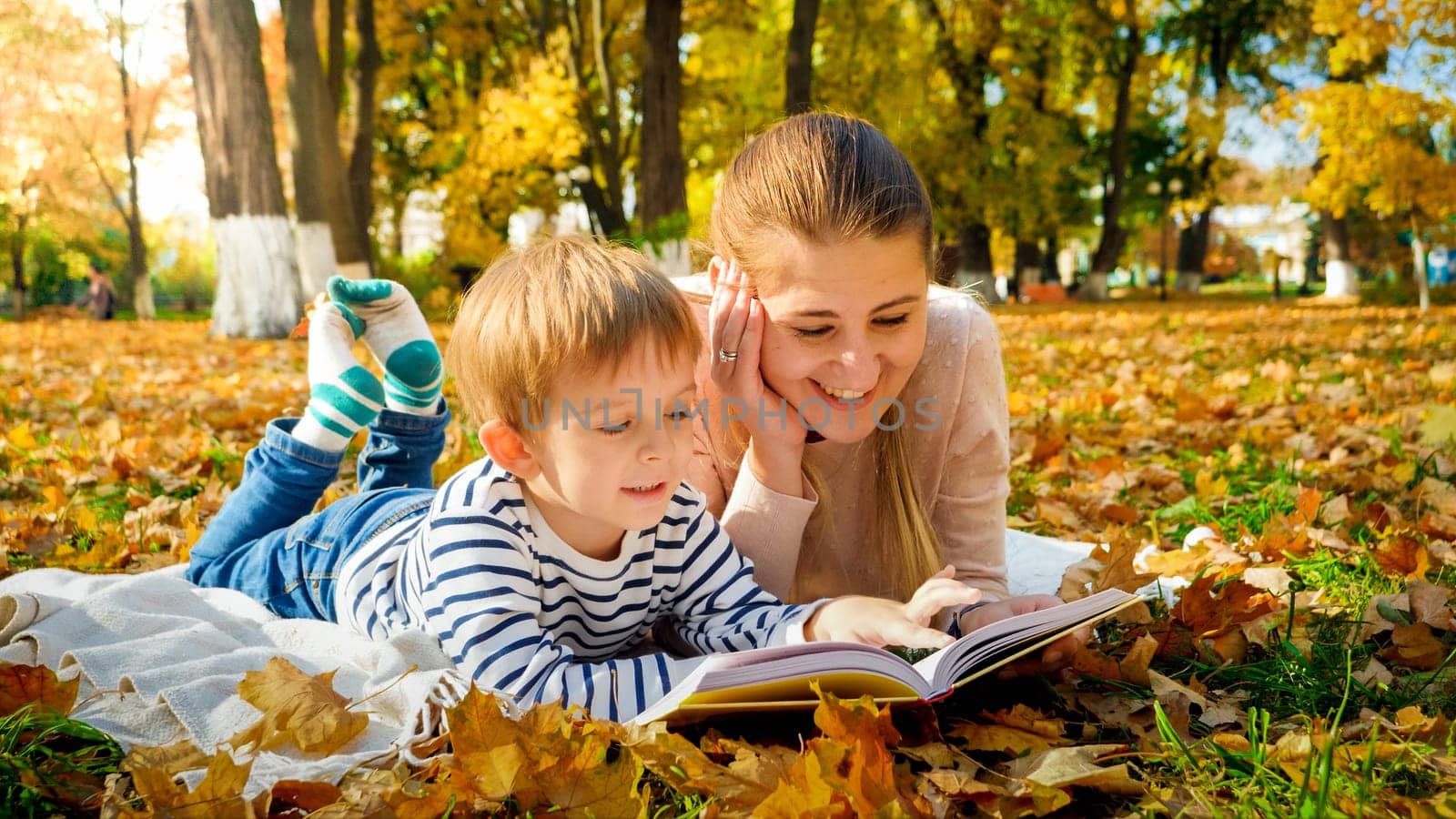 Portrait of happy smiling boy with mother reading book on picnic at autumn park.