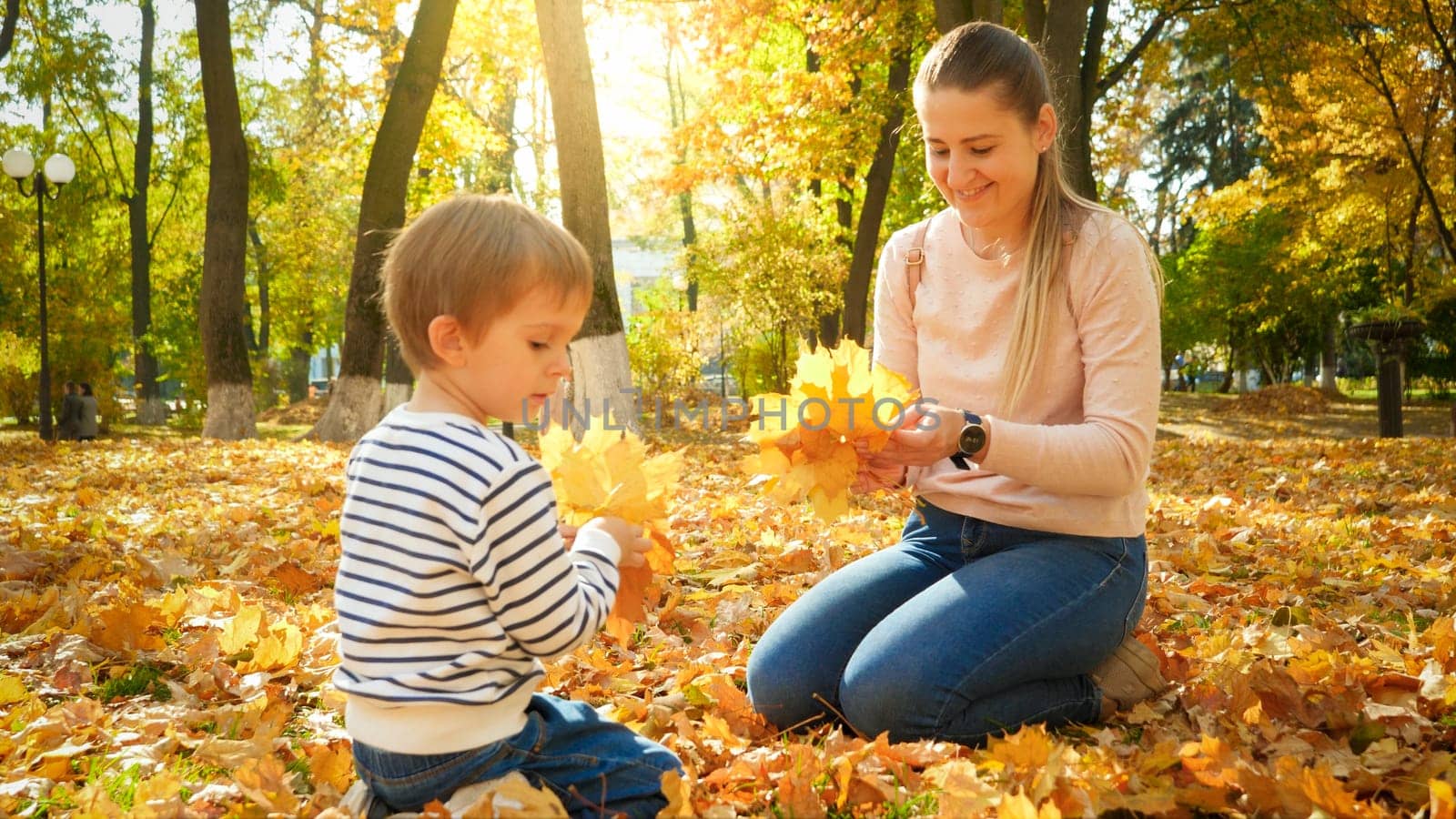 Portrait of beautiful smiling woman with little son picking up fallen yellow leaves in autumn park.