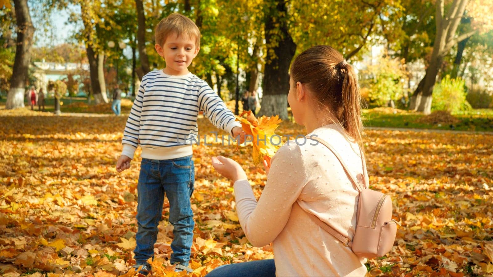 Happy smiling little boy with mother in autumn park covere with fallen yellow leaves.