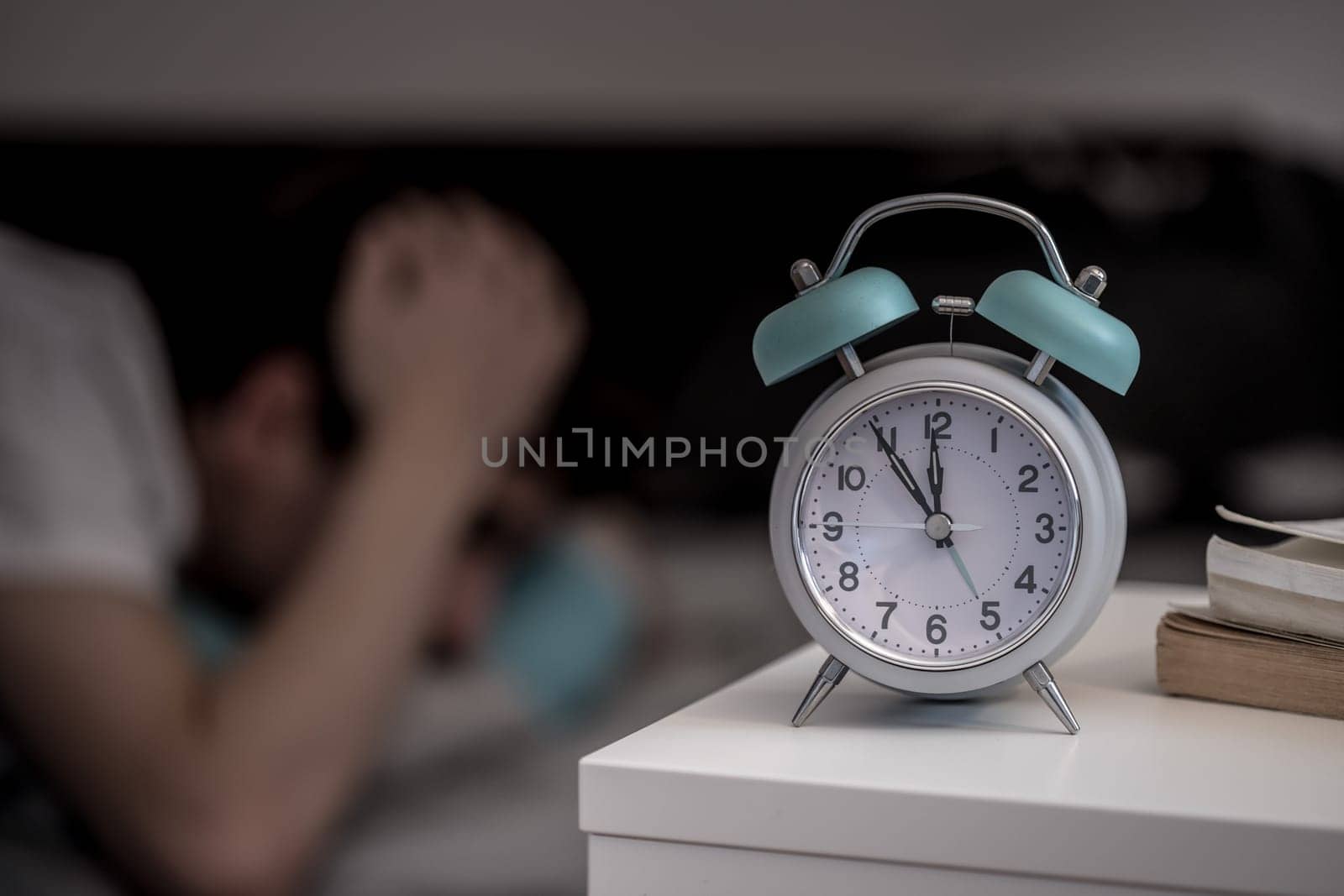 White alarm clock on the bedside table. Young tries to sleep in the background, insomnia
