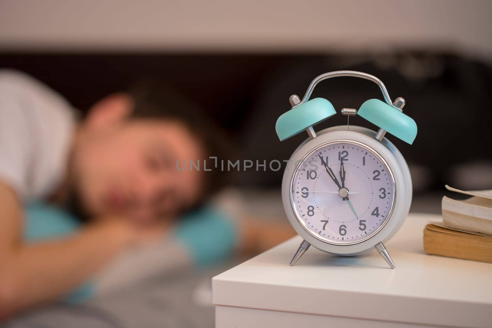 White alarm clock on the bedside table. Young man sleeps in the background.