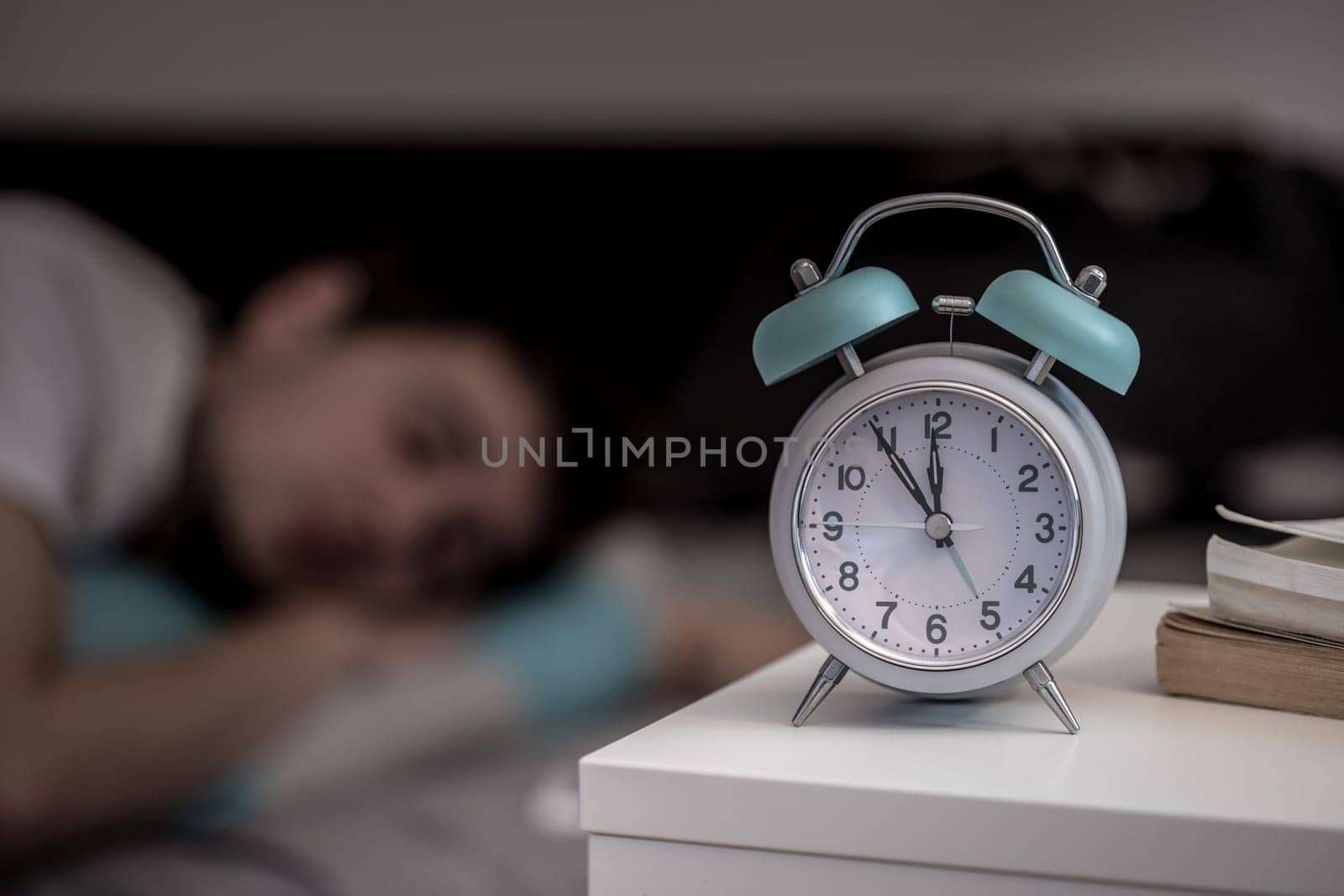 White alarm clock on the bedside table. Young man sleeps in the background.