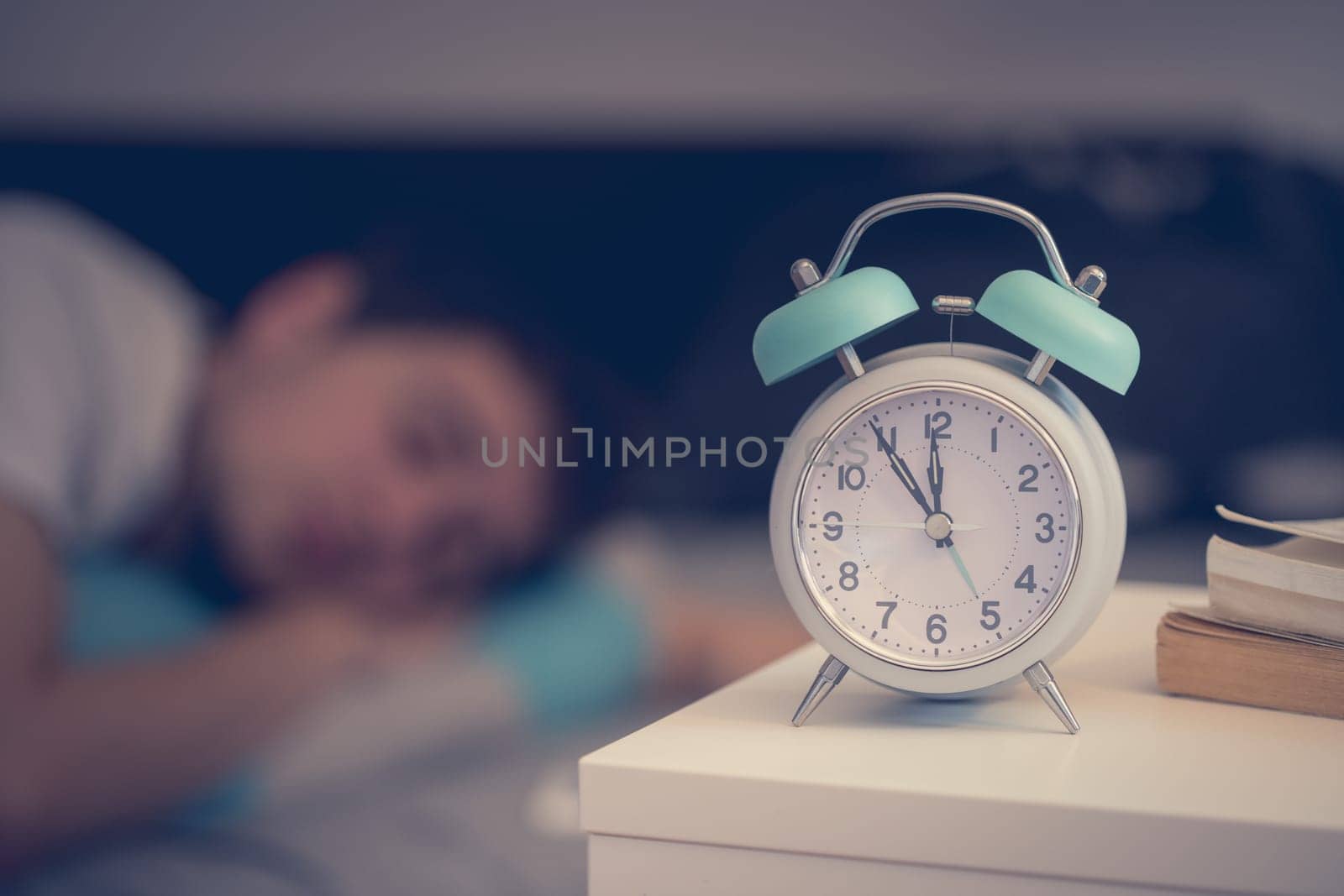 White alarm clock on the bedside table. Young man sleeps in the background.