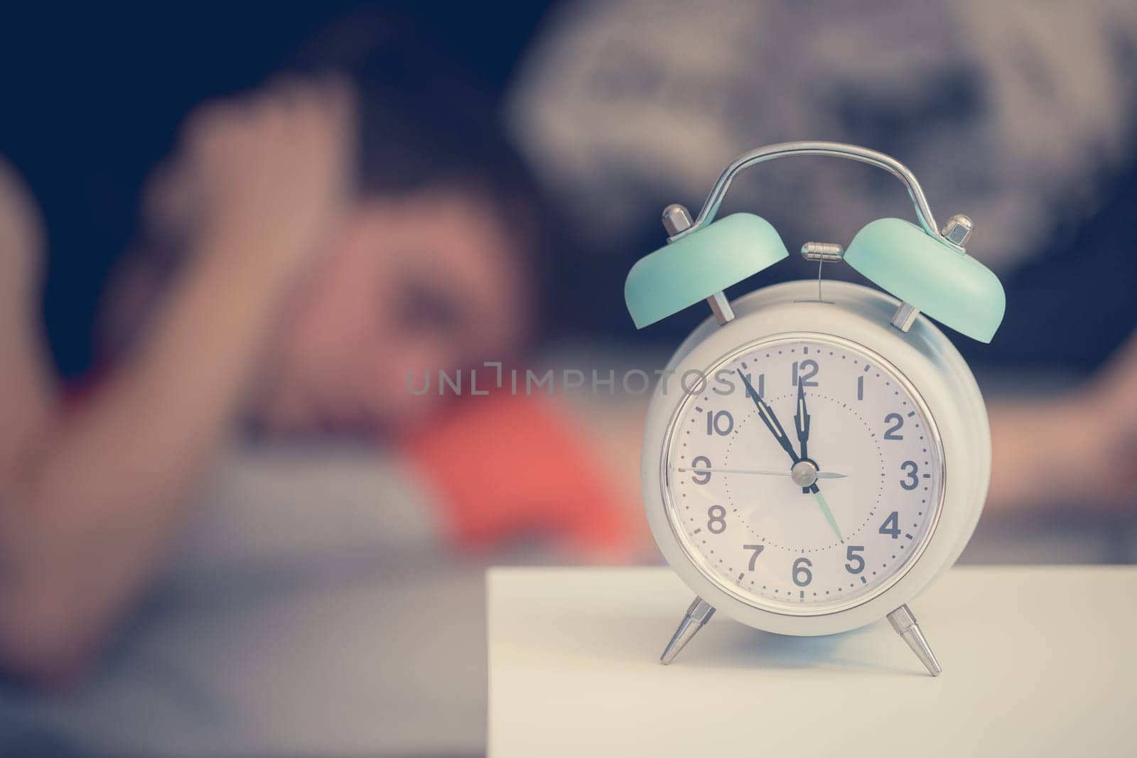 White alarm clock on the bedside table. Young tries to sleep in the background, insomnia