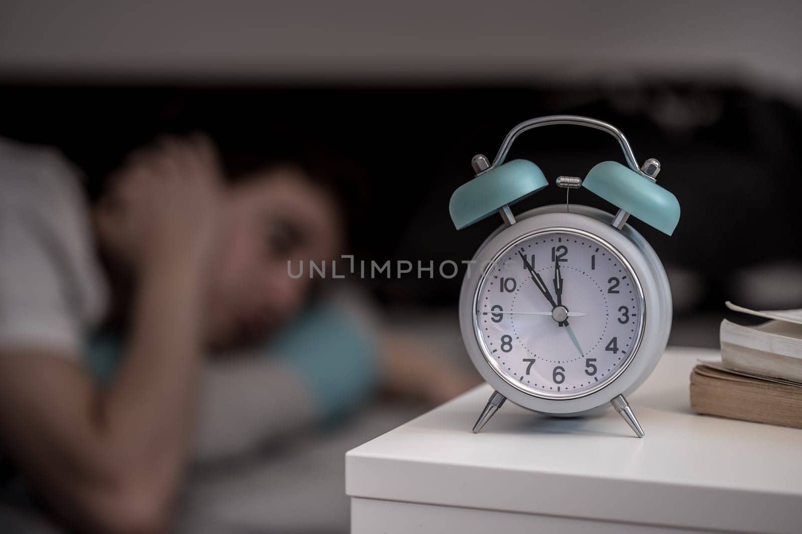 White alarm clock on the bedside table. Young tries to sleep in the background, insomnia